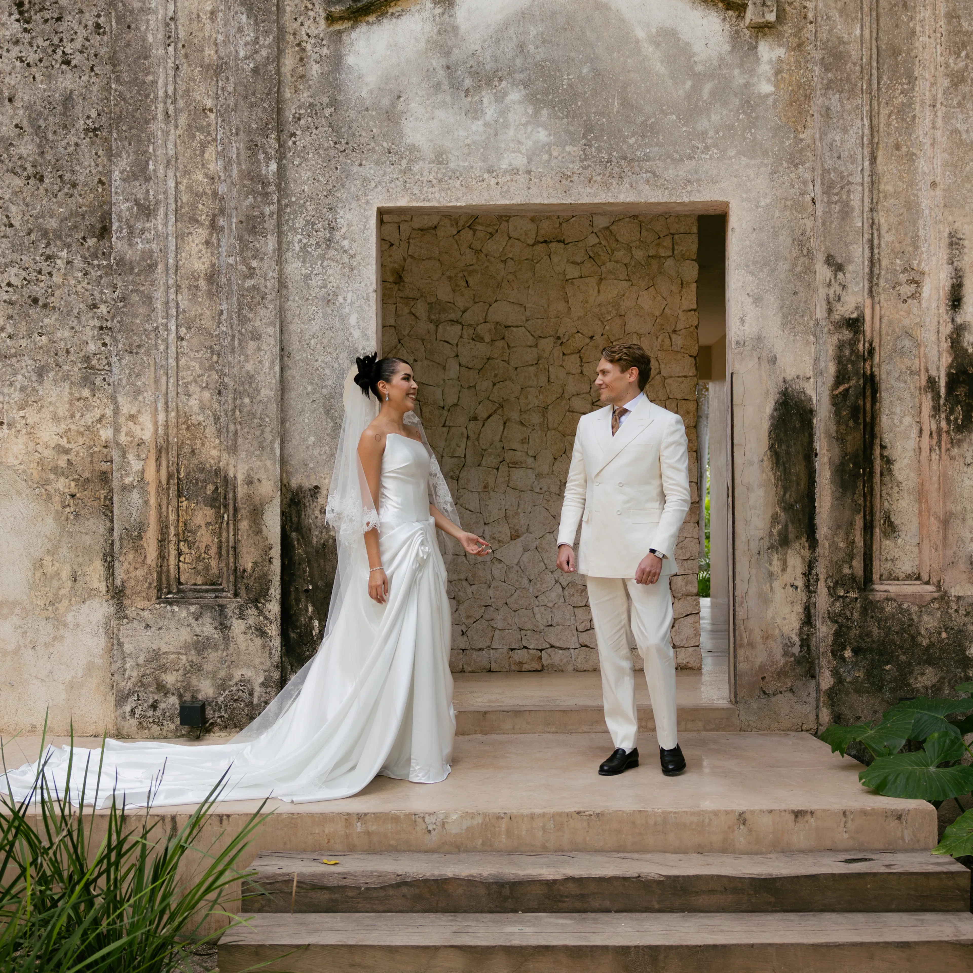 Los novios, elegantes vestidos de blanco, se encuentran en unos escalones de piedra, enmarcados por una pared desgastada y texturizada. La escena transmite una atmósfera romántica y atemporal.