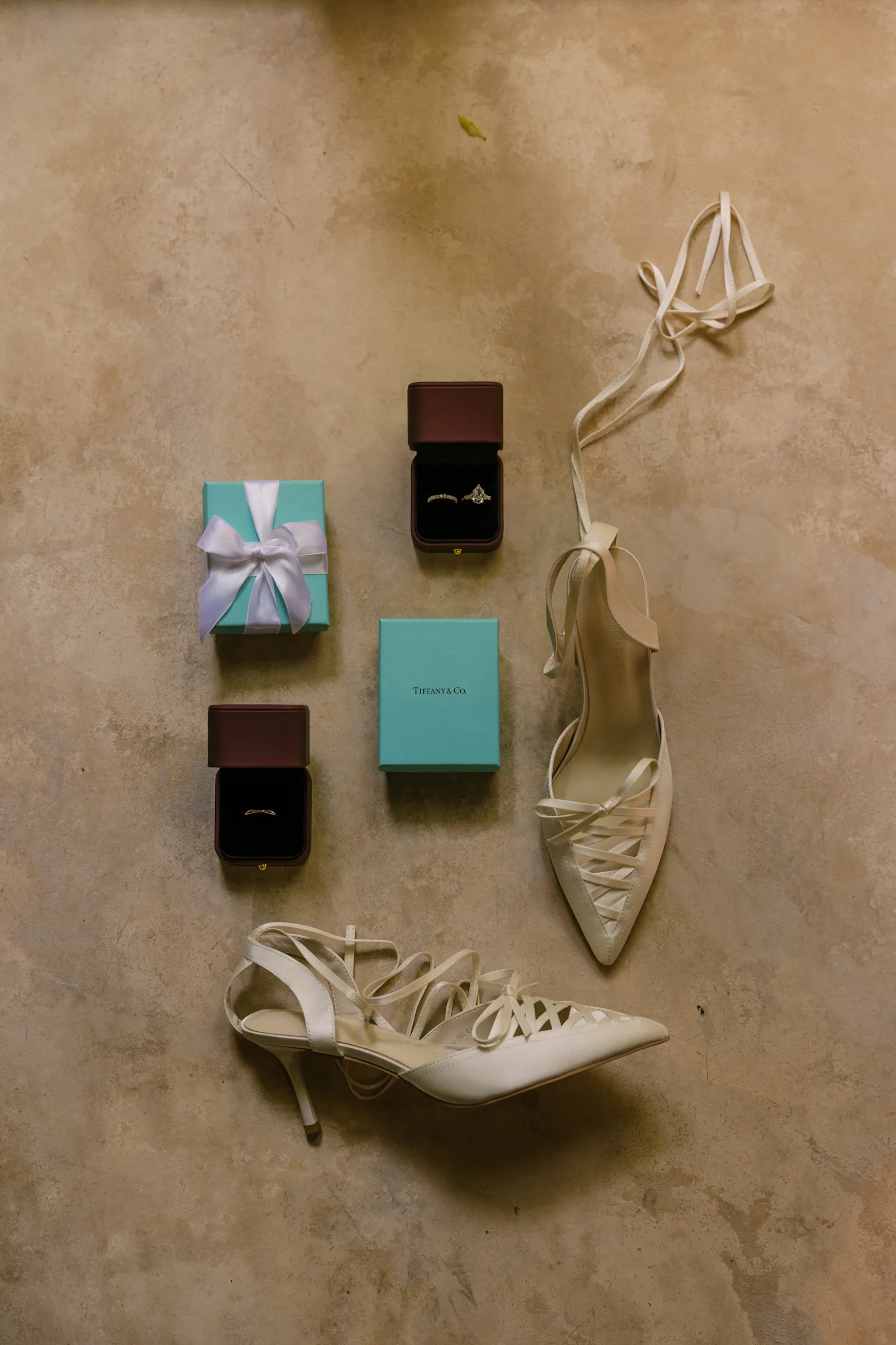 Elegantes tacones blancos con cordones, dos joyeros color verde azulado y dos cajas de anillos abiertas sobre una superficie beige, que transmiten una sensación de lujo y celebración.