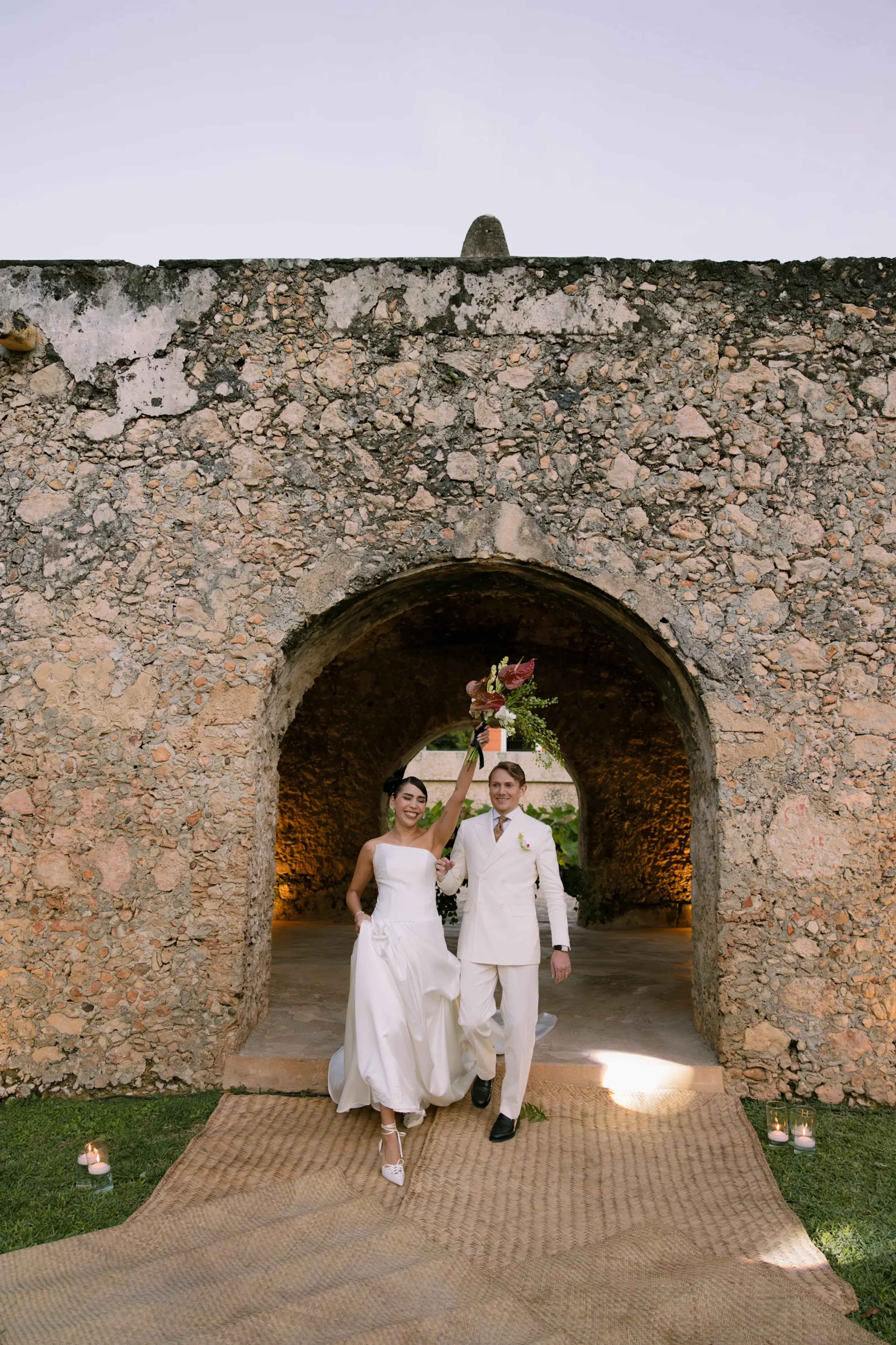 Los novios caminan alegremente por el arco de piedra, mientras ella alza el ramo triunfalmente. Ataviados con elegantes atuendos blancos, irradian felicidad y celebración.