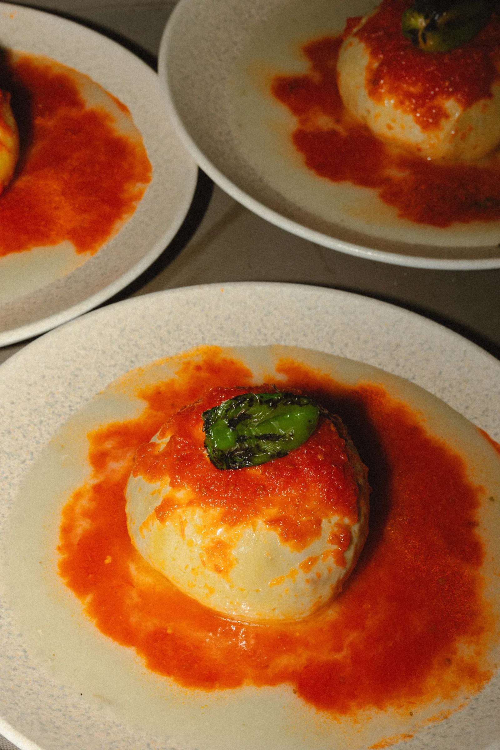 Una bola de masa redonda, cubierta con una salsa roja intensa y una rodaja de pimiento verde vibrante, se sirve en un plato blanco. La imagen transmite una sensación rica y apetitosa.