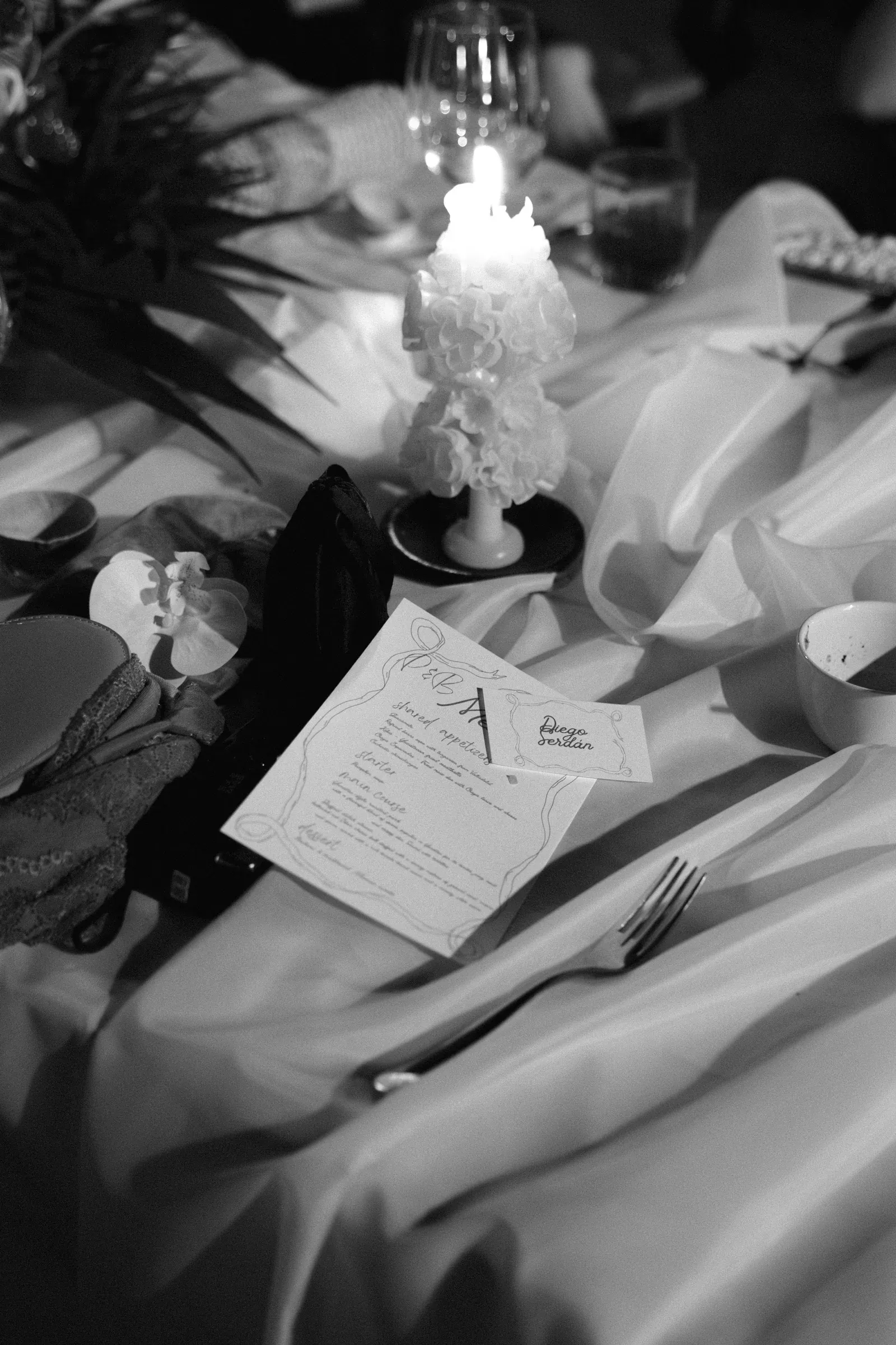 Imagen en blanco y negro de una mesa elegantemente puesta con una vela texturizada, un menú y una orquídea. La iluminación suave crea un ambiente cálido e íntimo.