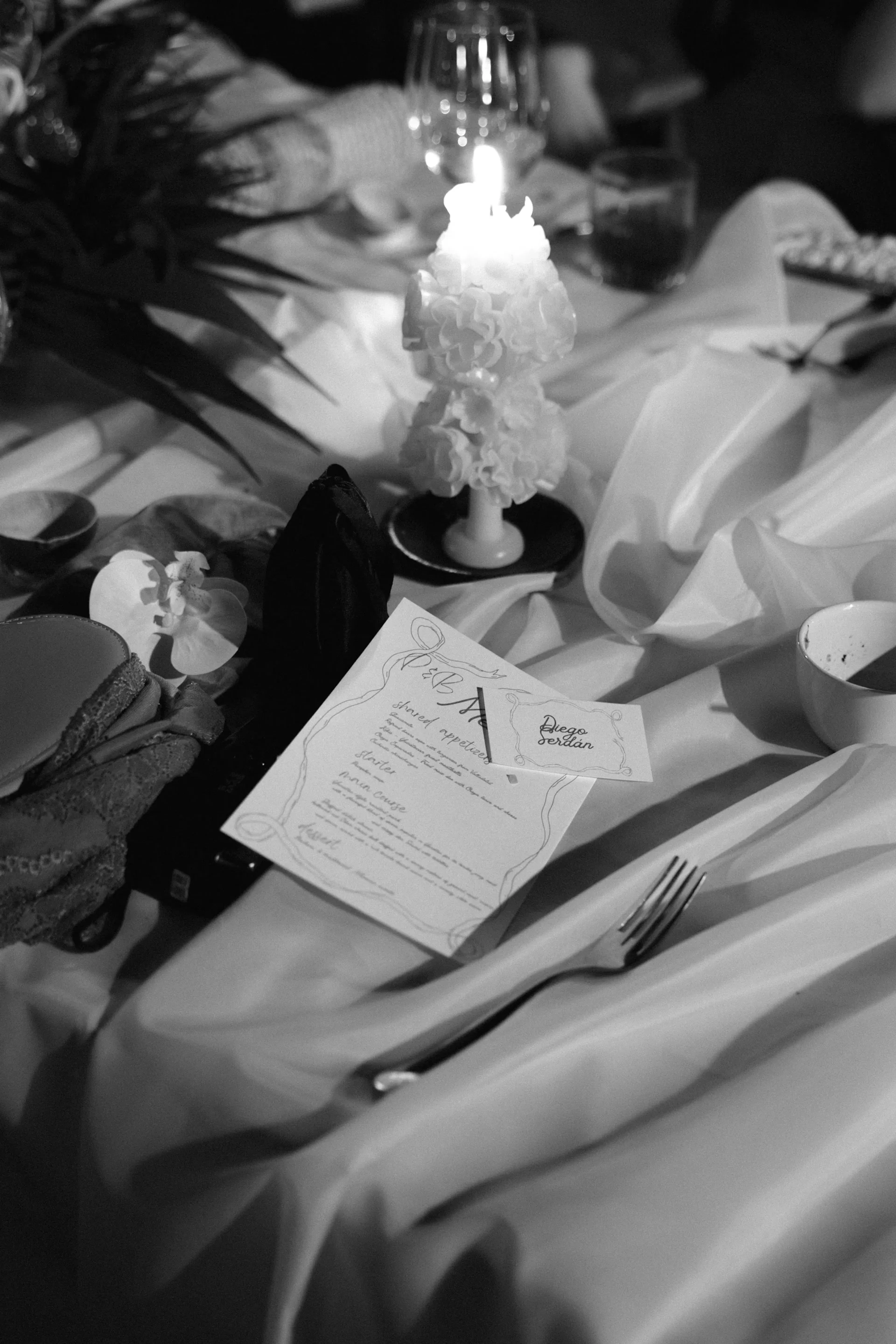 Imagen en blanco y negro de una mesa elegantemente puesta con una vela texturizada, un menú y una orquídea. La iluminación suave crea un ambiente cálido e íntimo.