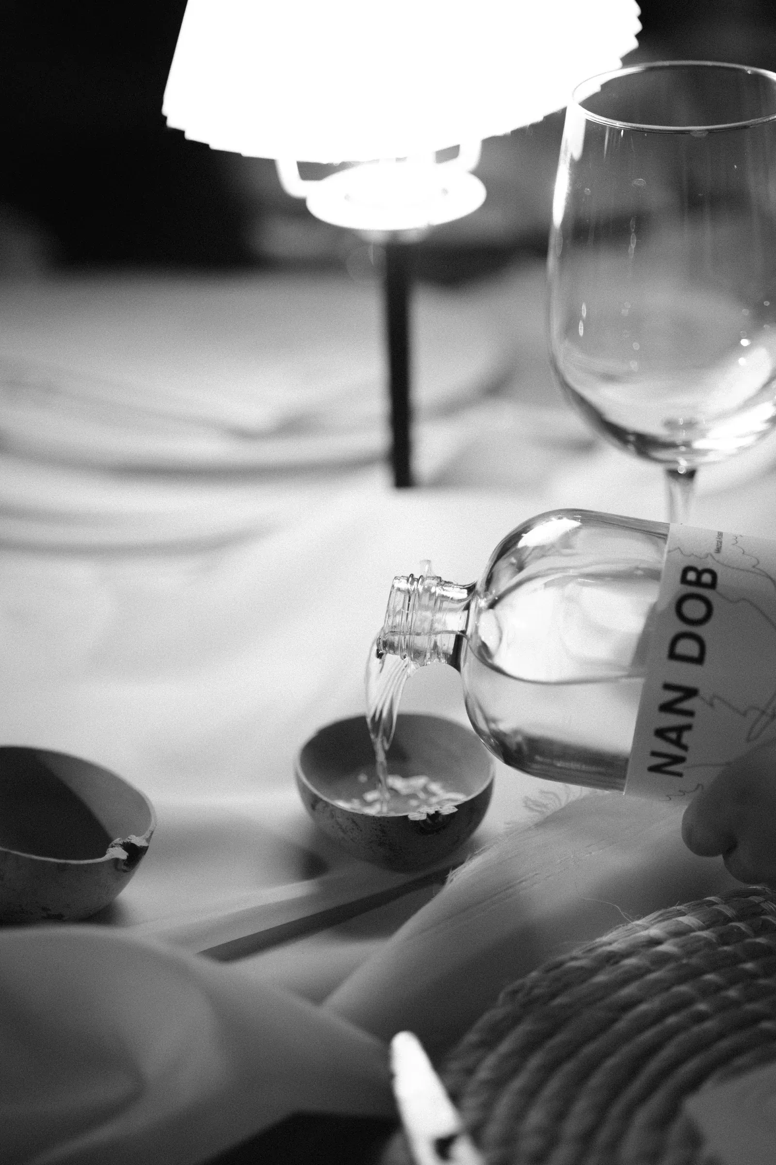 Imagen en blanco y negro de una mesa elegantemente puesta con una botella sirviendo bebida en un recipiente. La iluminación suave crea un ambiente cálido e íntimo.