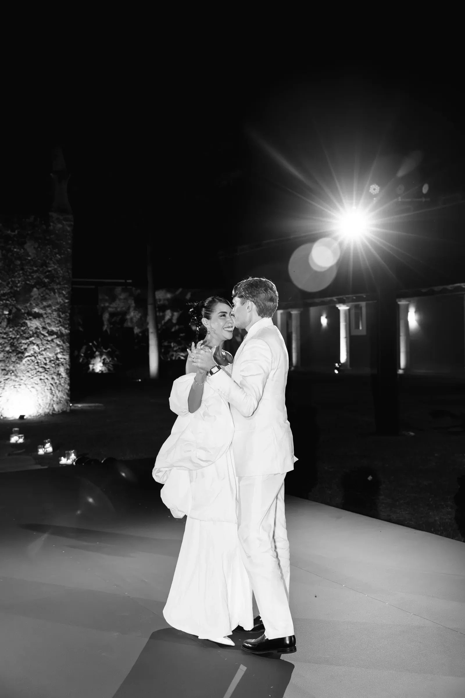 Una pareja baila alegremente con su atuendo nupcial bajo un foco brillante en un oscuro escenario nocturno al aire libre. La escena rezuma romance y celebración.