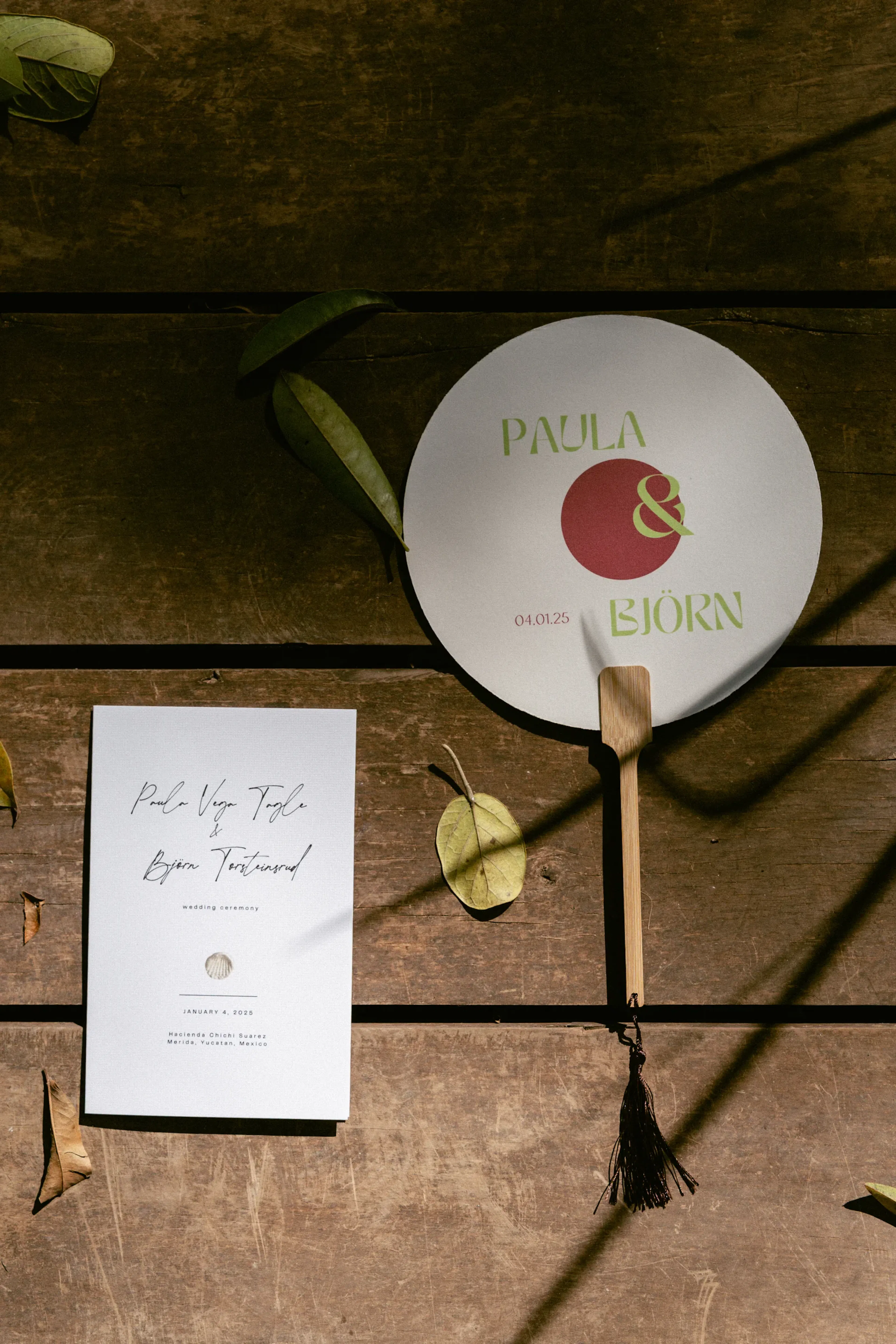 Una invitación de boda sobre una superficie de madera, acompañada de un abanico redondo con el texto "Paula & Björn" en verde. La luz del sol proyecta sombras de hojas a su alrededor.