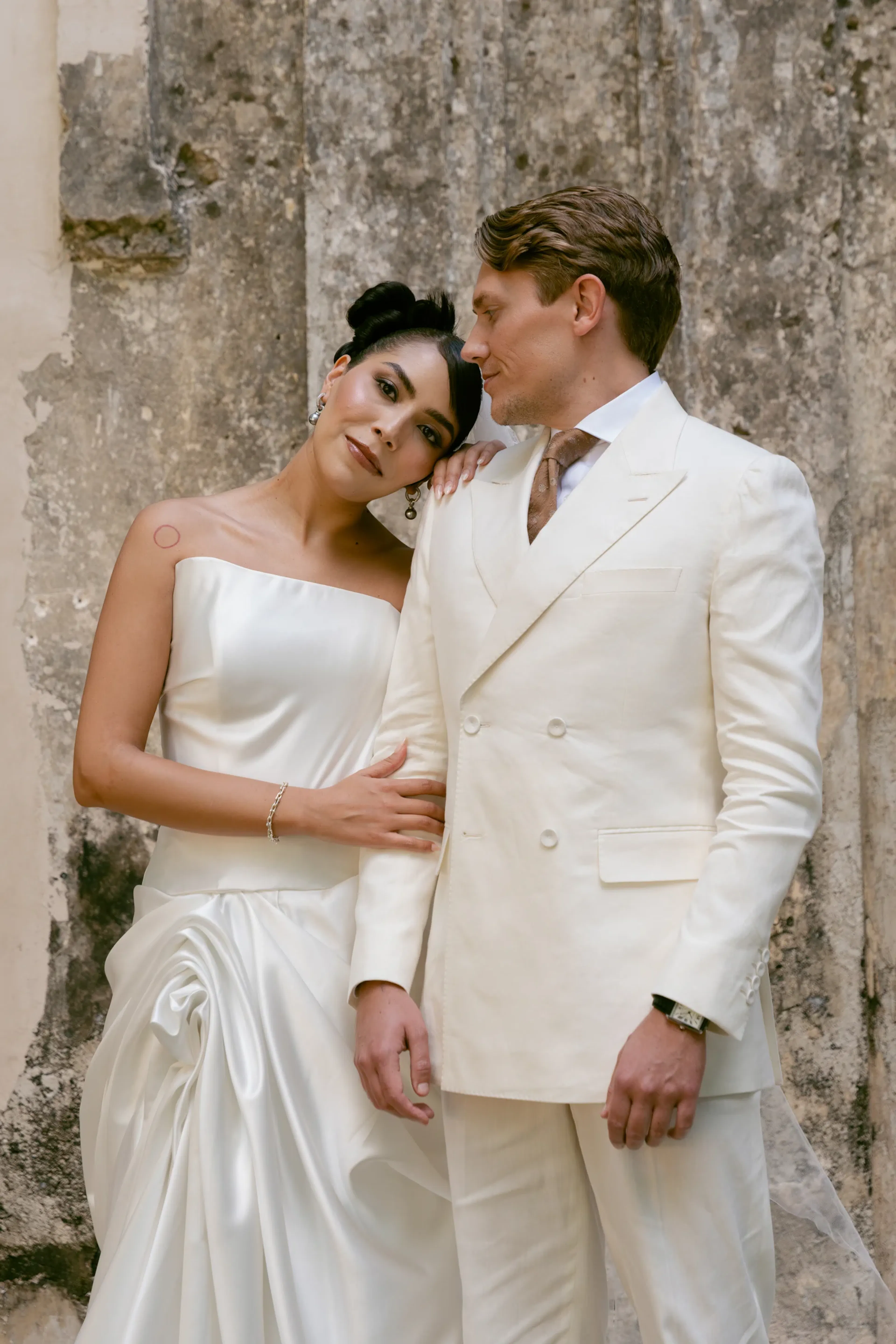 Una pareja, vestida elegantemente de blanco, se encuentra de pie frente a un muro de piedra texturizada. La mujer se apoya cariñosamente en el hombro del hombre, irradiando alegría y ternura.