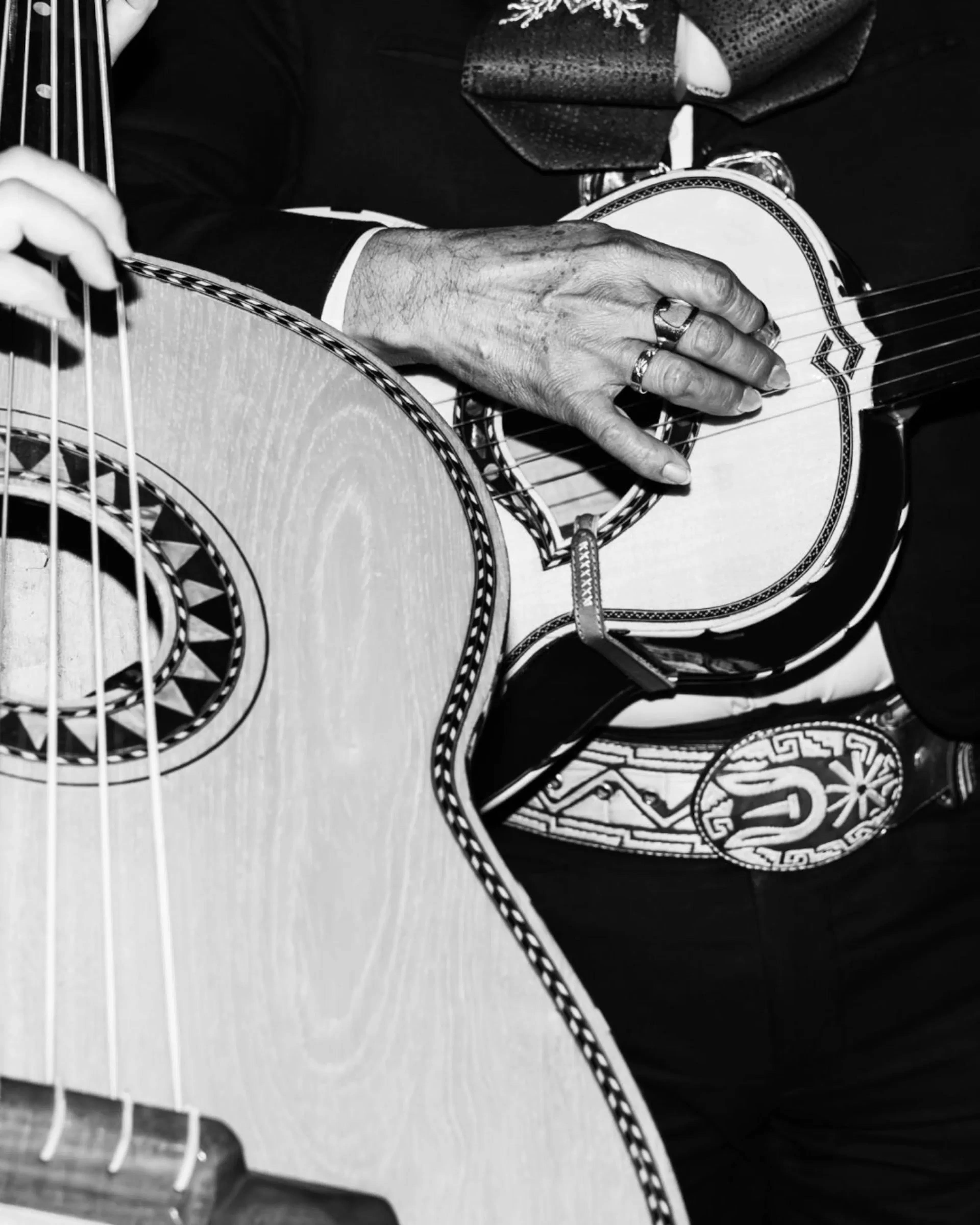 Primer plano de las manos de un músico tocando la guitarra. La imagen en blanco y negro muestra intrincados patrones en la guitarra y el cinturón adornado del músico.