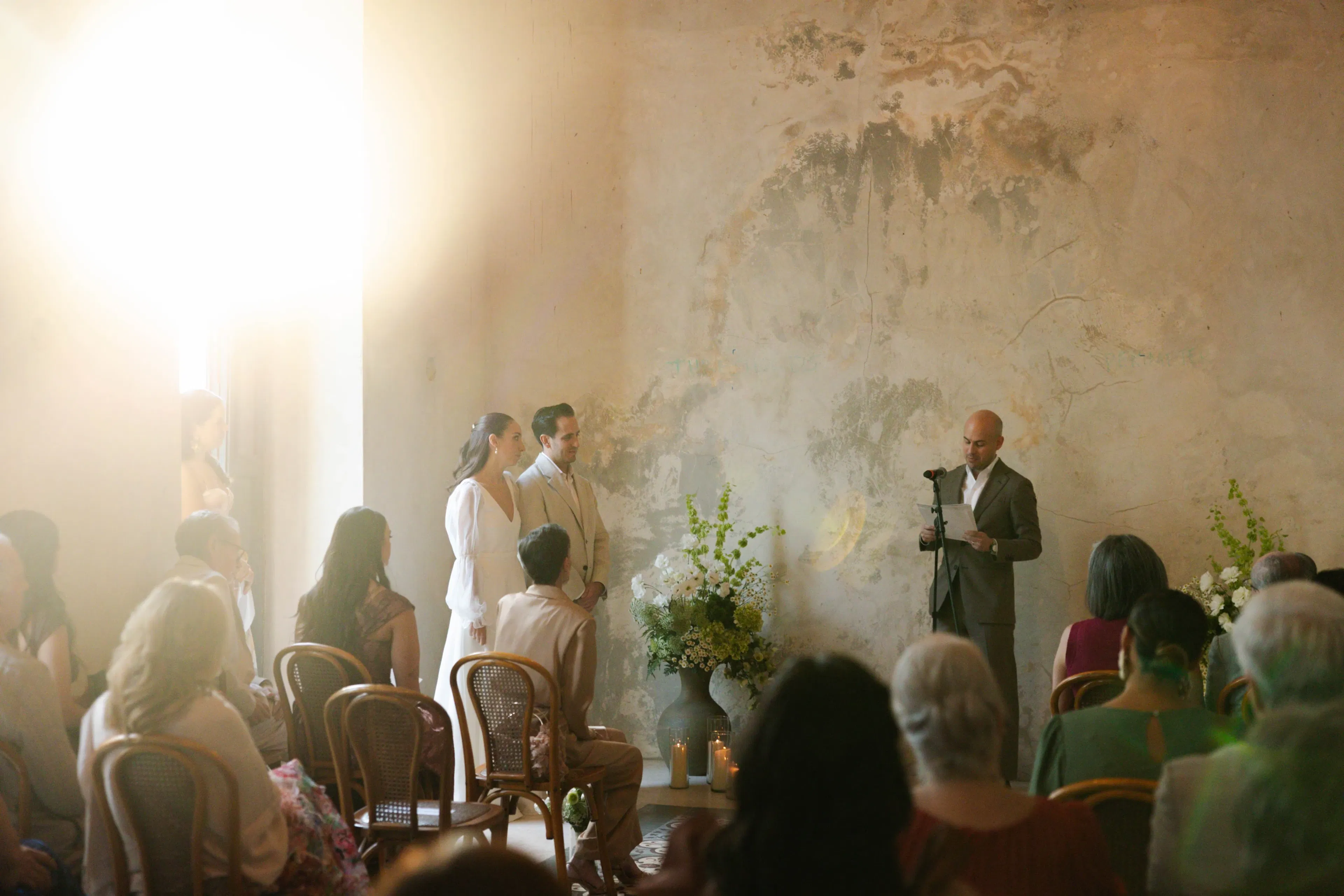 Una ceremonia de boda serena en una sala rústica y soleada. Una pareja está de pie ante el oficiante leyendo. Los invitados se sientan en sillas de madera, bajo una suave luz y rodeados de flores.