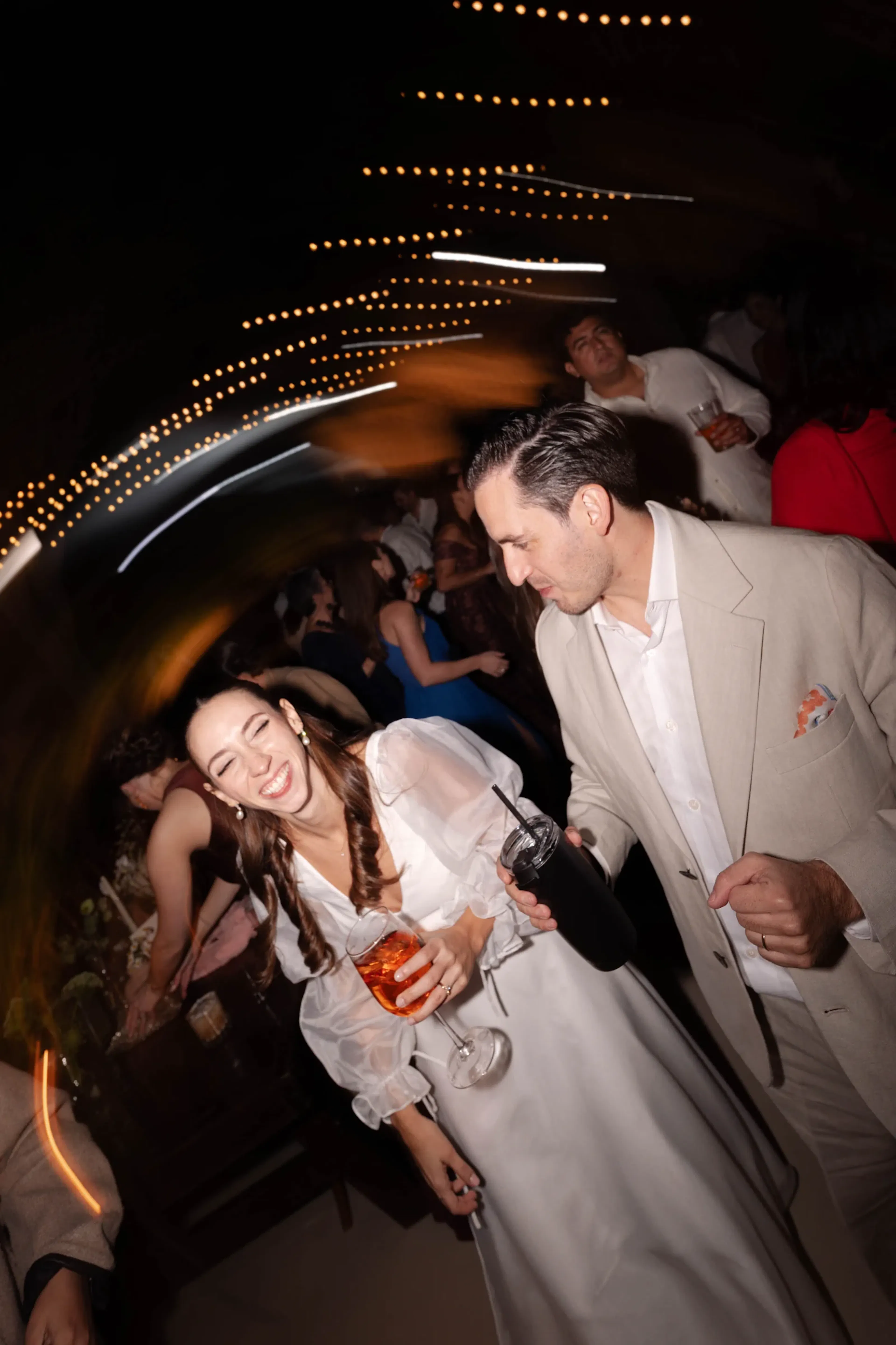 Una pareja alegre baila en una fiesta. La mujer, vestida de blanco, ríe mientras sostiene una copa. El hombre, vestido de beige, la mira. Las luces se arremolinan en el cielo.