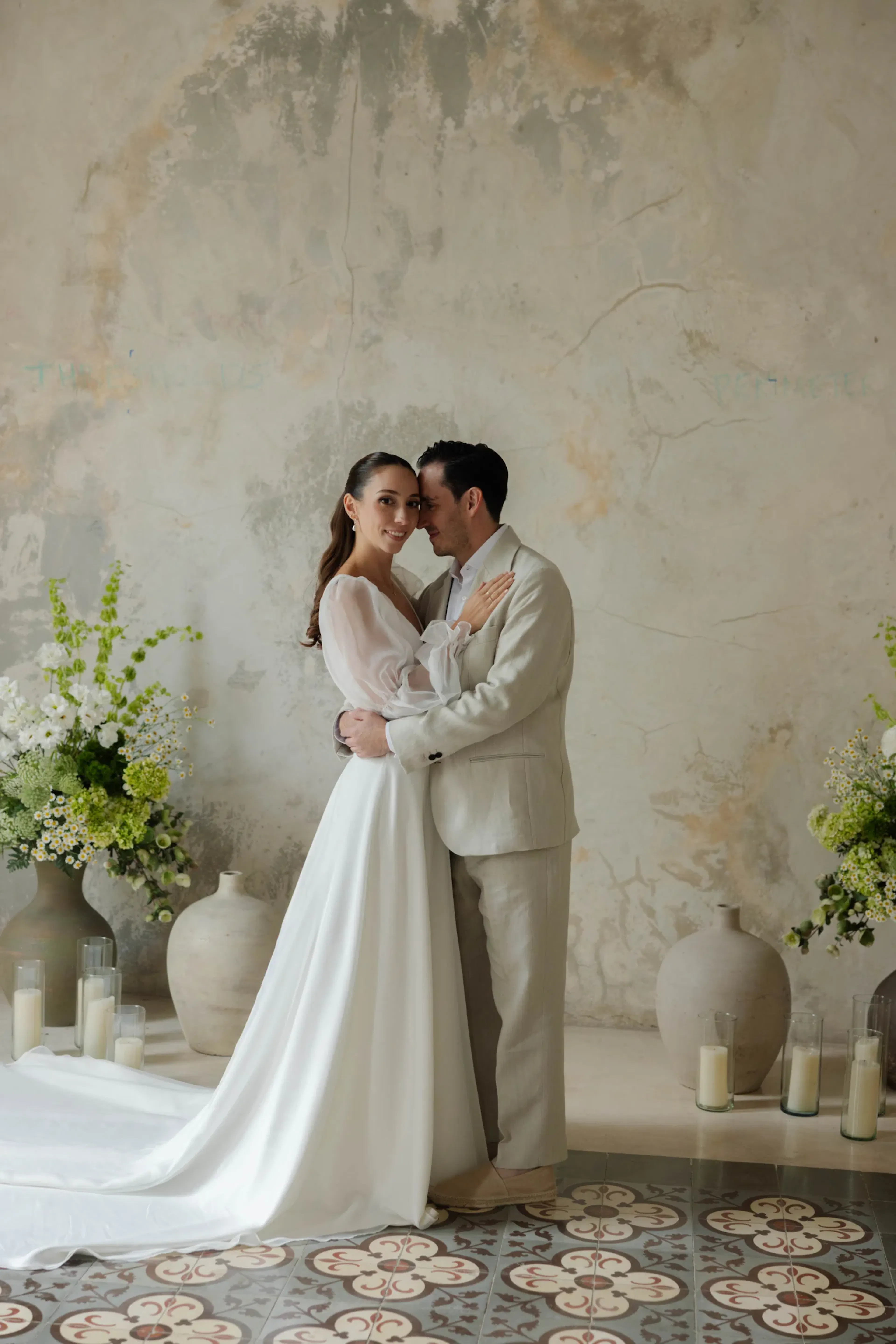 Una pareja vestida elegantemente se abraza contra una pared texturizada, rodeada de arreglos florales verdes y velas, transmitiendo romance y serenidad.