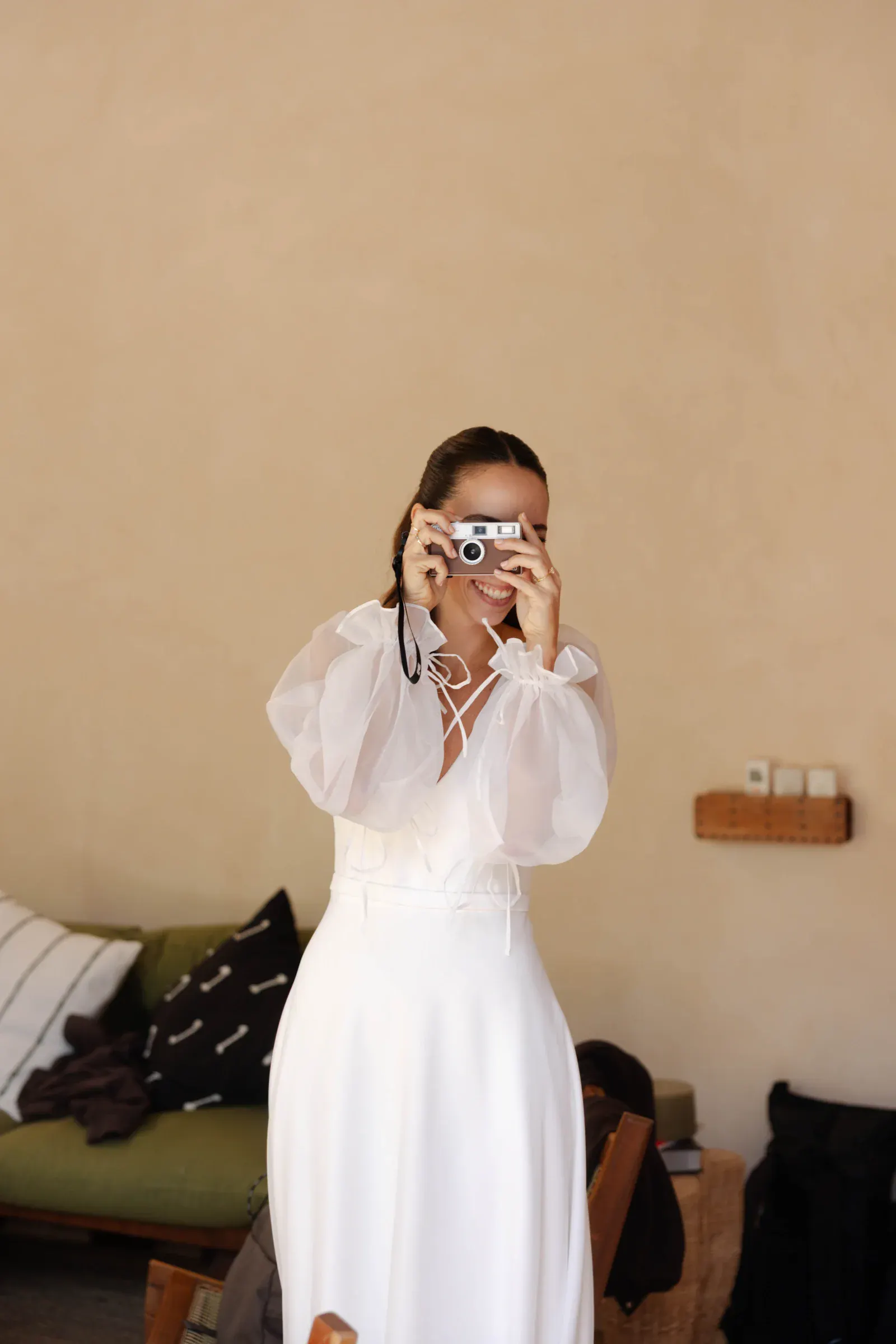Una persona con un vestido blanco y mangas transparentes sonríe mientras sostiene una cámara. Se encuentra en una habitación cálidamente iluminada con un sofá verde y cojines al fondo.