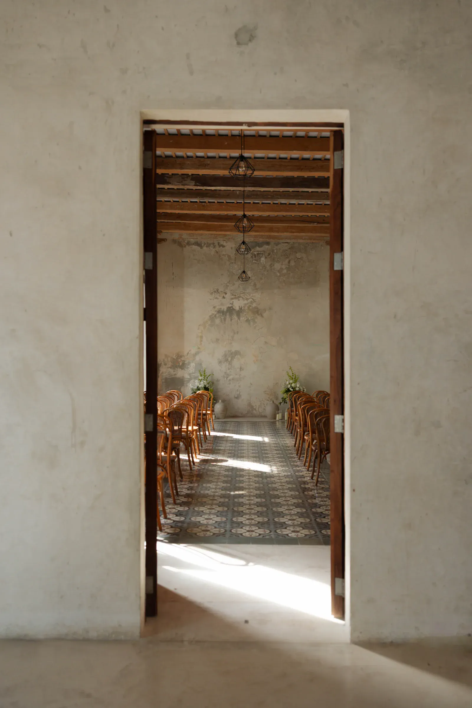 Al mirar a través de una puerta, se descubre una habitación rústica con sillas de madera alineadas sobre un suelo de baldosas estampadas. La luz tenue crea un ambiente sereno.