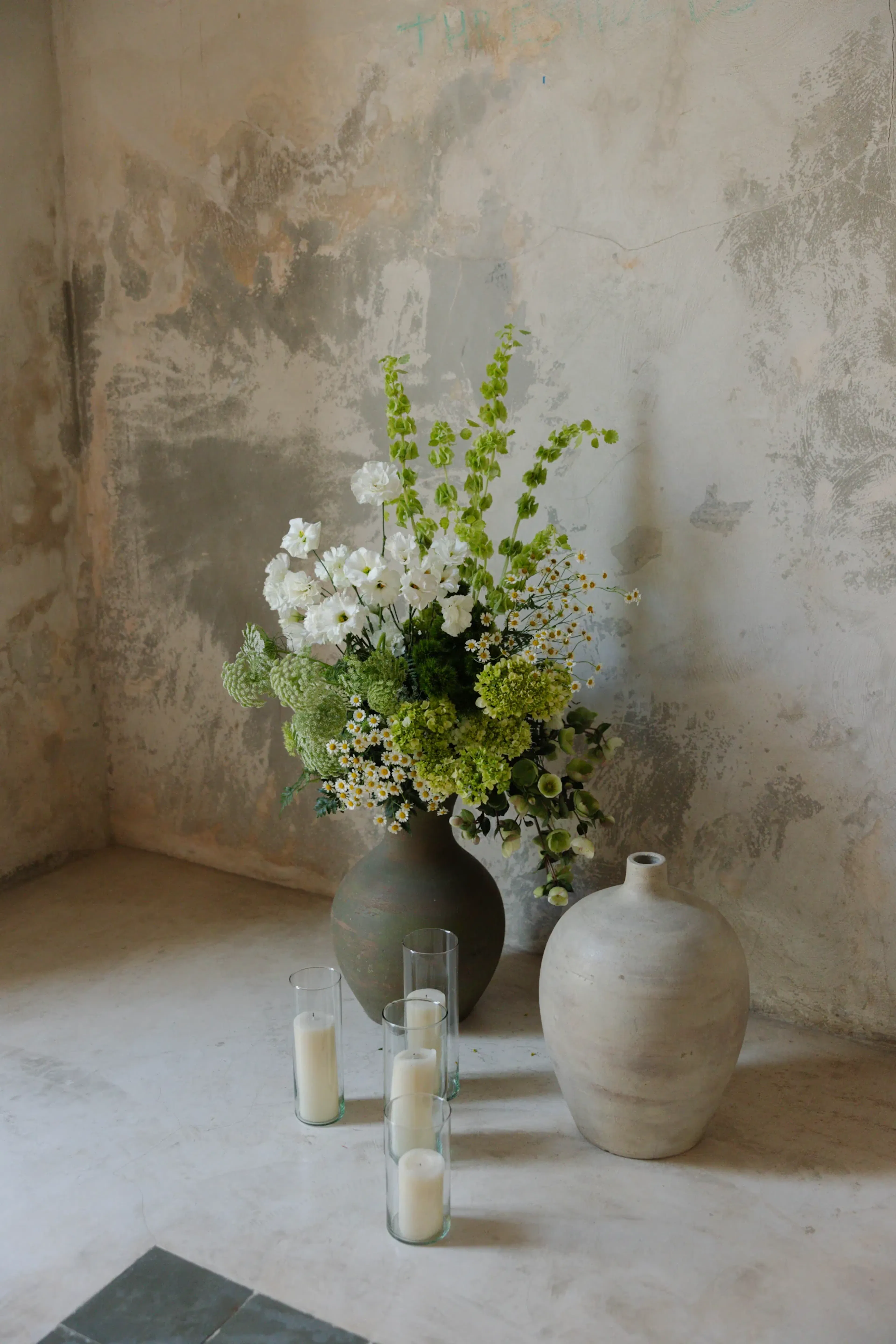 Una escena rústica con un ramo de flores blancas y verdes en un gran jarrón de barro, junto a una maceta redonda de barro. Cuatro velas altas de cristal descansan en el suelo, contra una pared texturizada y desgastada. El ambiente es tranquilo y sereno.