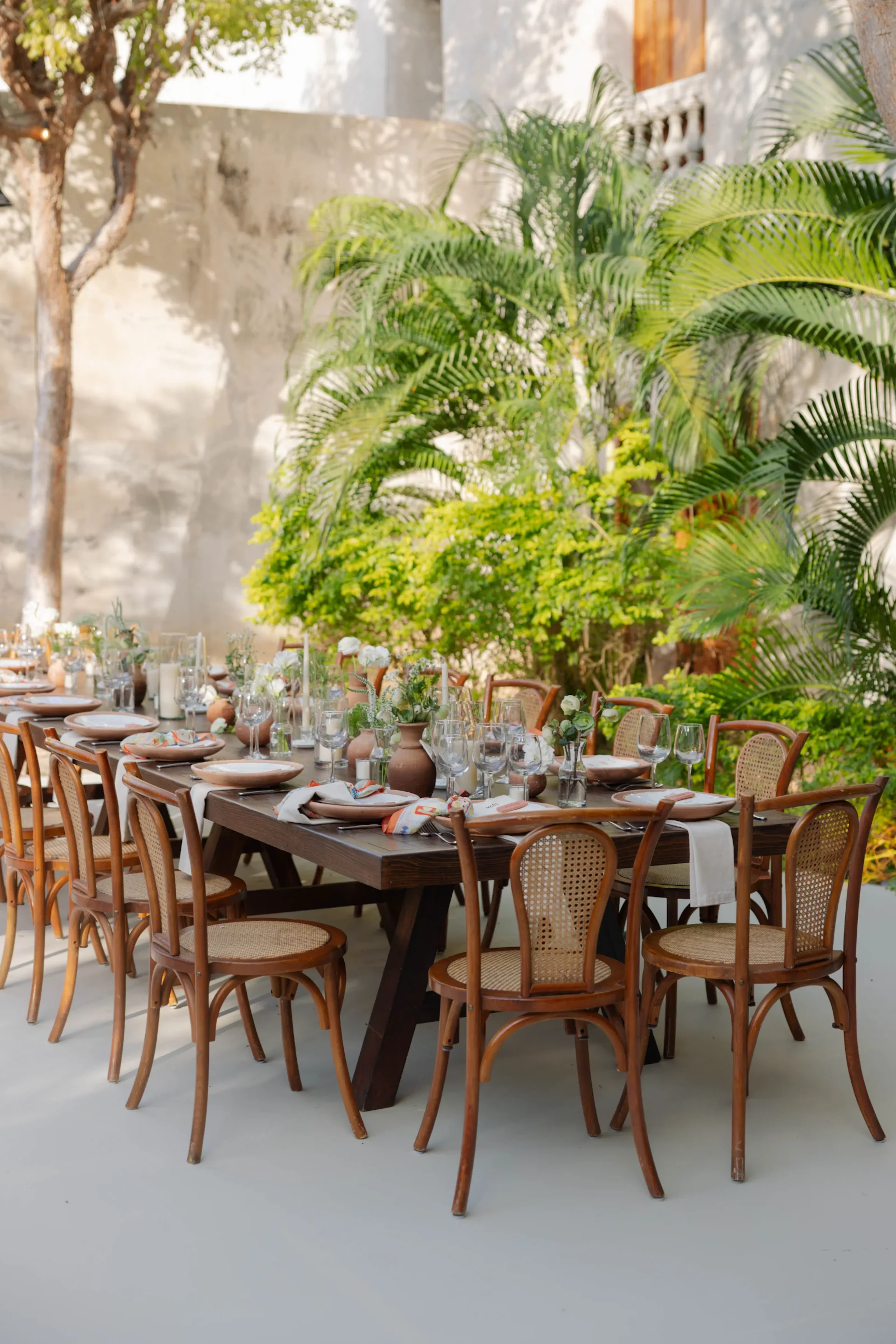 Un elegante comedor al aire libre cuenta con una mesa larga con sillas de madera, platos, vasos y jarrones con flores. Una exuberante vegetación rodea el espacio, creando un ambiente sereno y acogedor.
