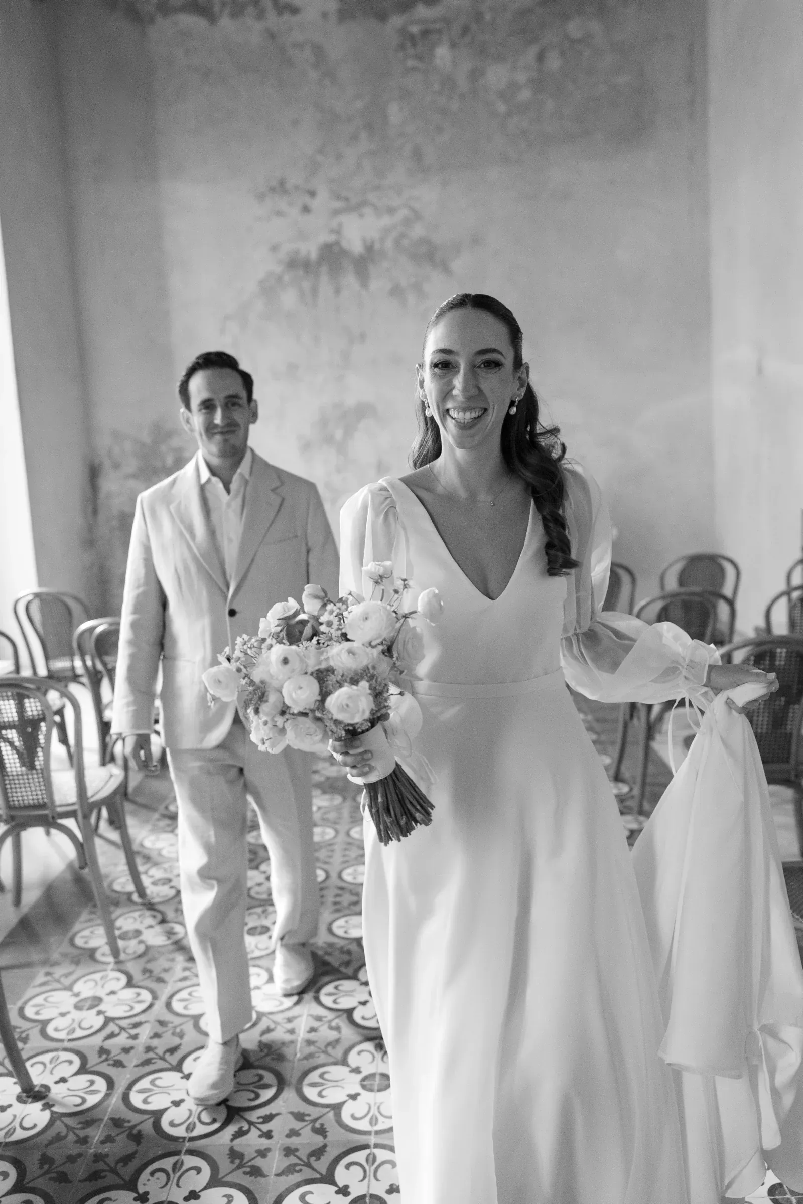 La novia, con un elegante vestido blanco, sostiene un ramo de flores y sonríe con alegría. El novio, con un traje ligero, la sigue. El suelo de baldosas vintage y la iluminación tenue crean un ambiente romántico.