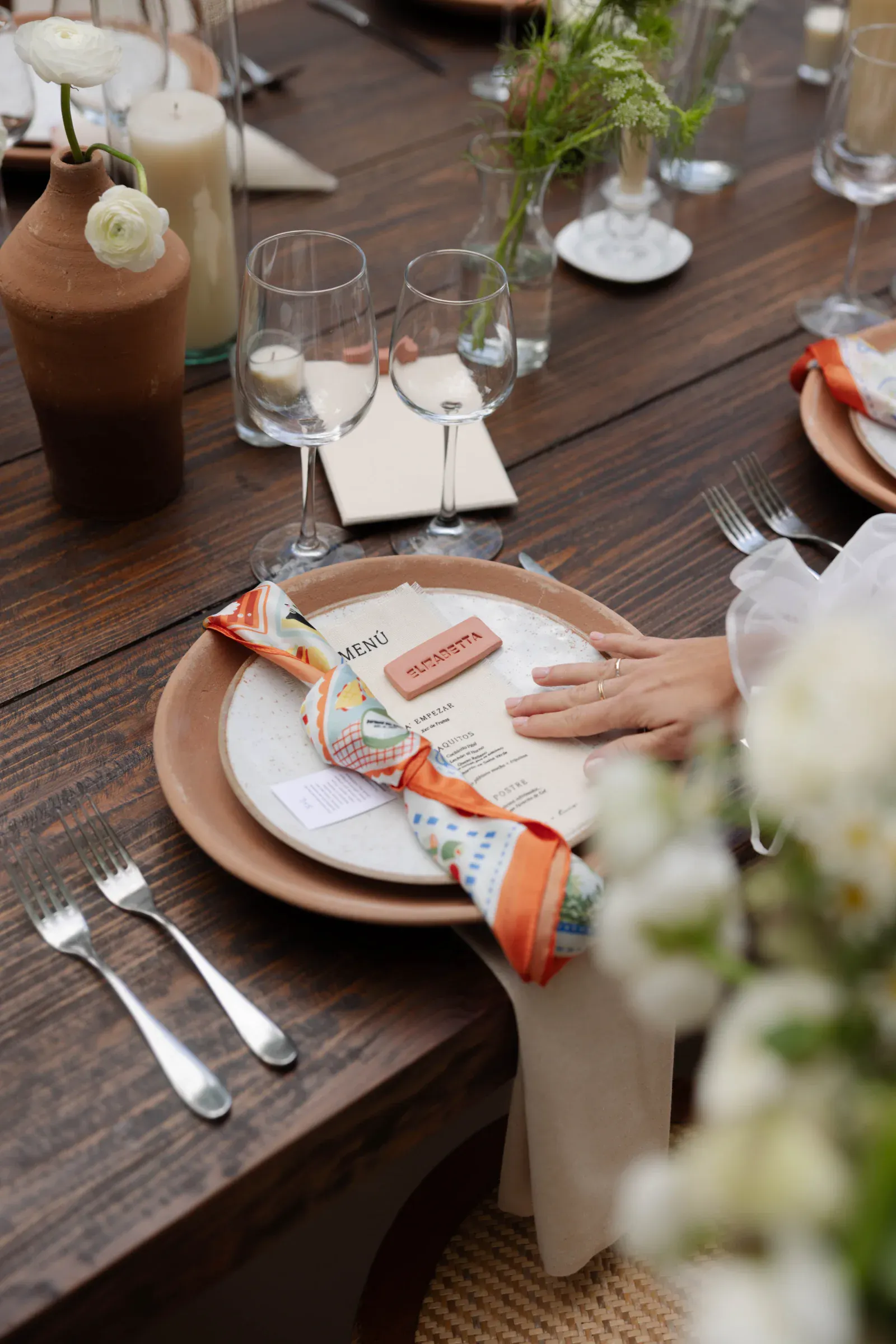 Elegante mesa de estilo rústico, con un plato marrón con un menú decorativo, una servilleta colorida, copas de vino y un centro de mesa floral. Una mano descansa suavemente cerca.