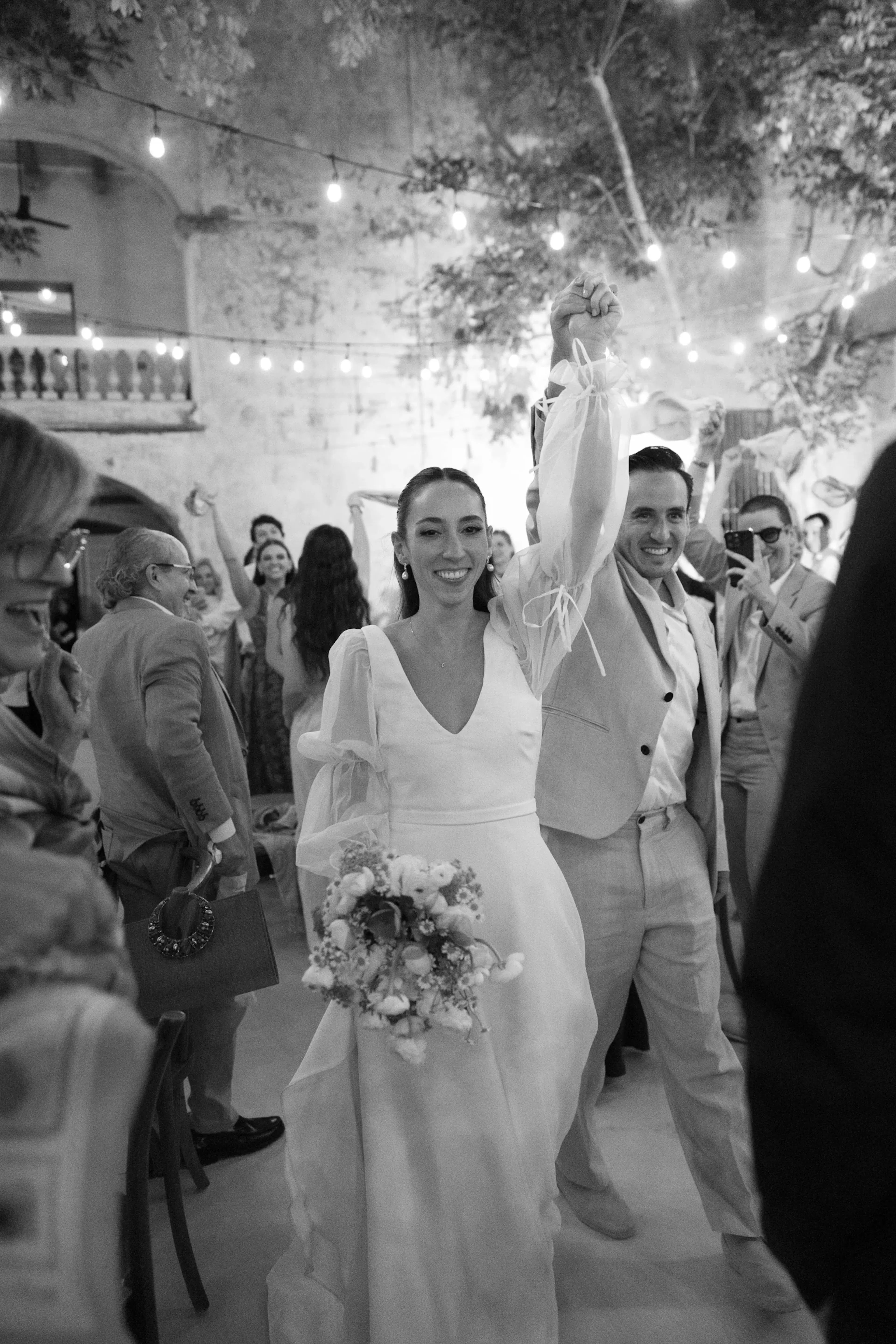 Una alegre pareja con traje de novia camina entre la multitud, la novia con un ramo en la mano. Las guirnaldas de luces crean un ambiente festivo. Abundan las sonrisas.