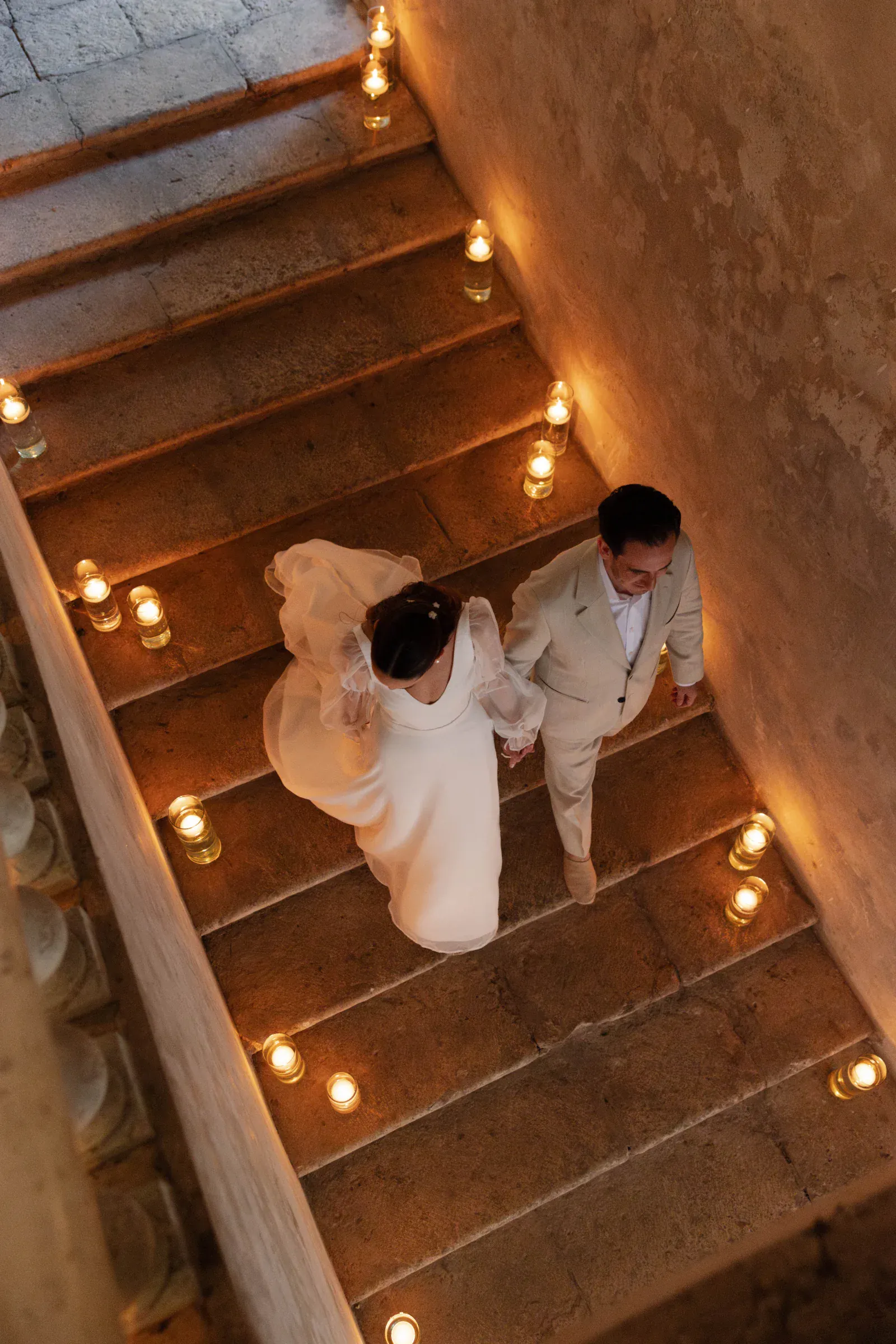 Una pareja vestida formal camina de la mano por unos escalones de piedra bordeados de velas brillantes, creando un ambiente cálido y romántico.