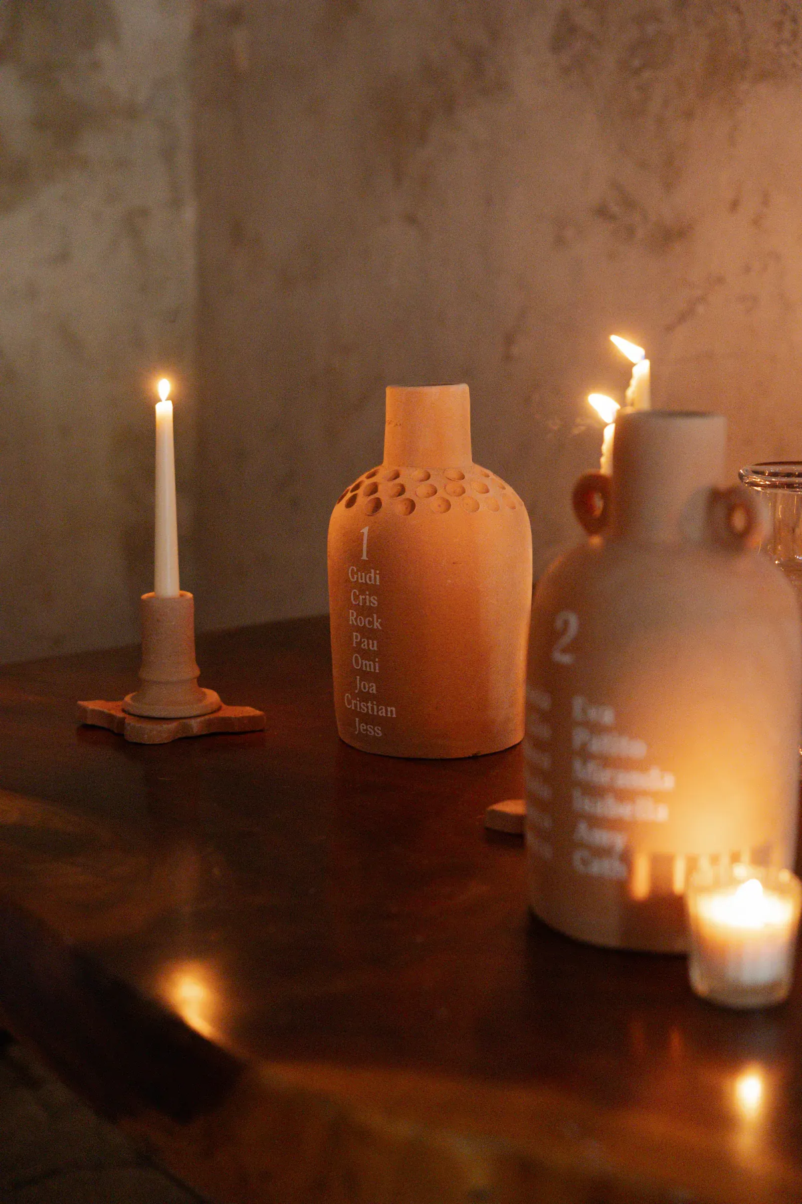 Dos jarrones de cerámica con nombres inscritos se encuentran sobre una mesa de madera tenuemente iluminada, acompañados de velas brillantes, creando una atmósfera cálida y serena.