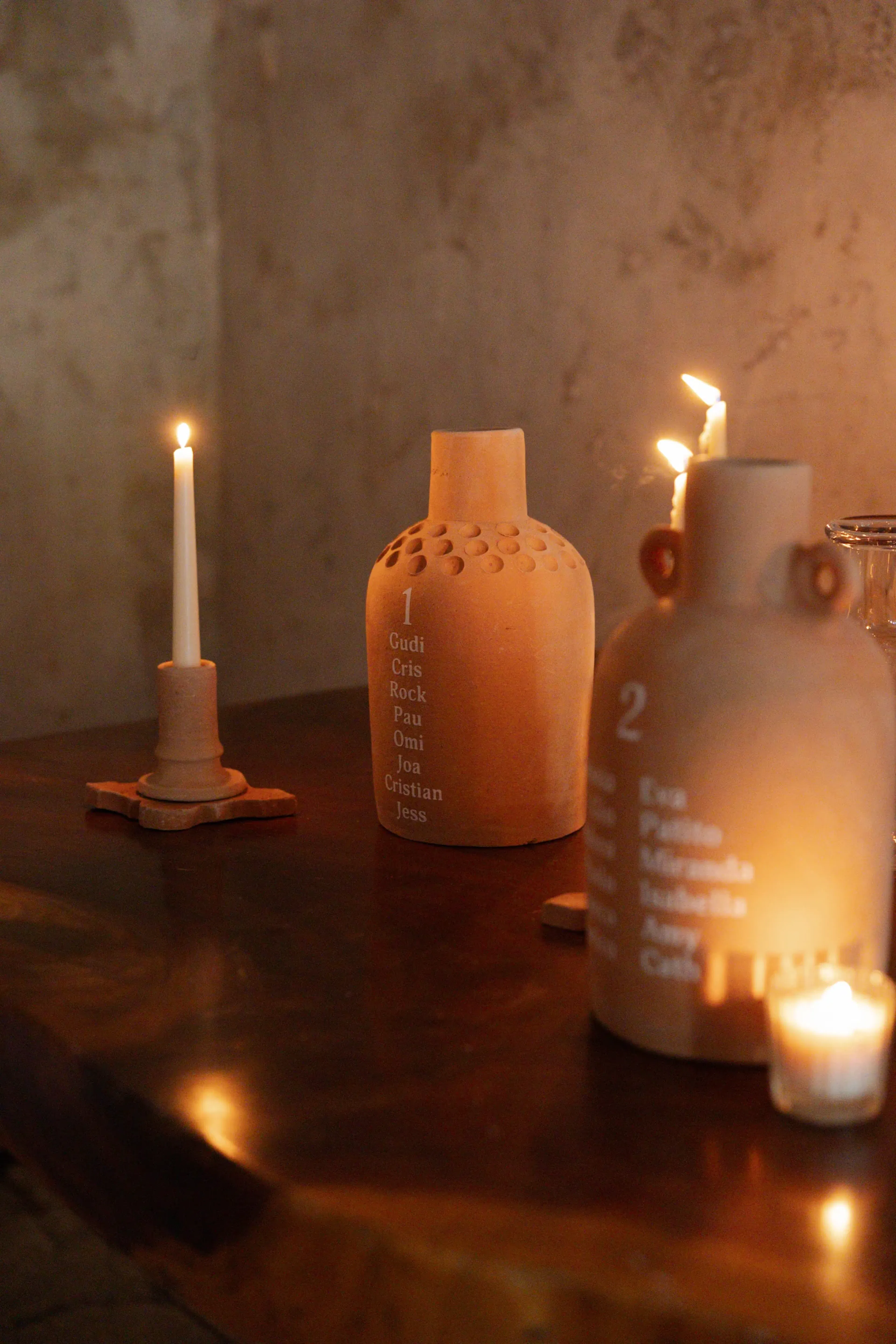 Dos jarrones de cerámica con nombres inscritos se encuentran sobre una mesa de madera tenuemente iluminada, acompañados de velas brillantes, creando una atmósfera cálida y serena.