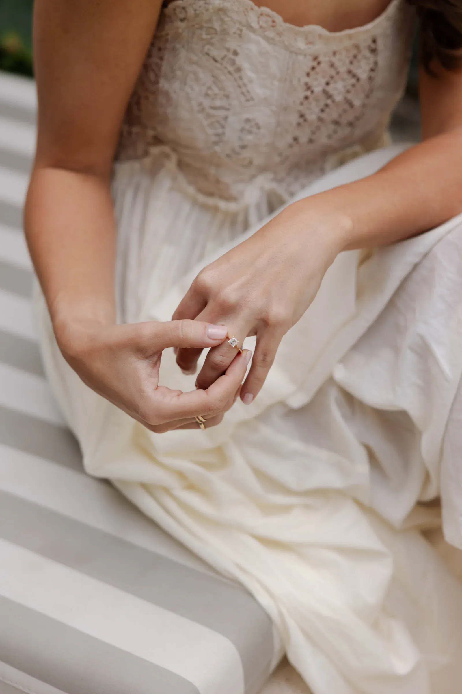 Una mujer con un vestido de encaje ajusta un anillo en su dedo mientras está sentada sobre cojines de rayas grises y blancas, transmitiendo un estado de ánimo sereno y contemplativo.
