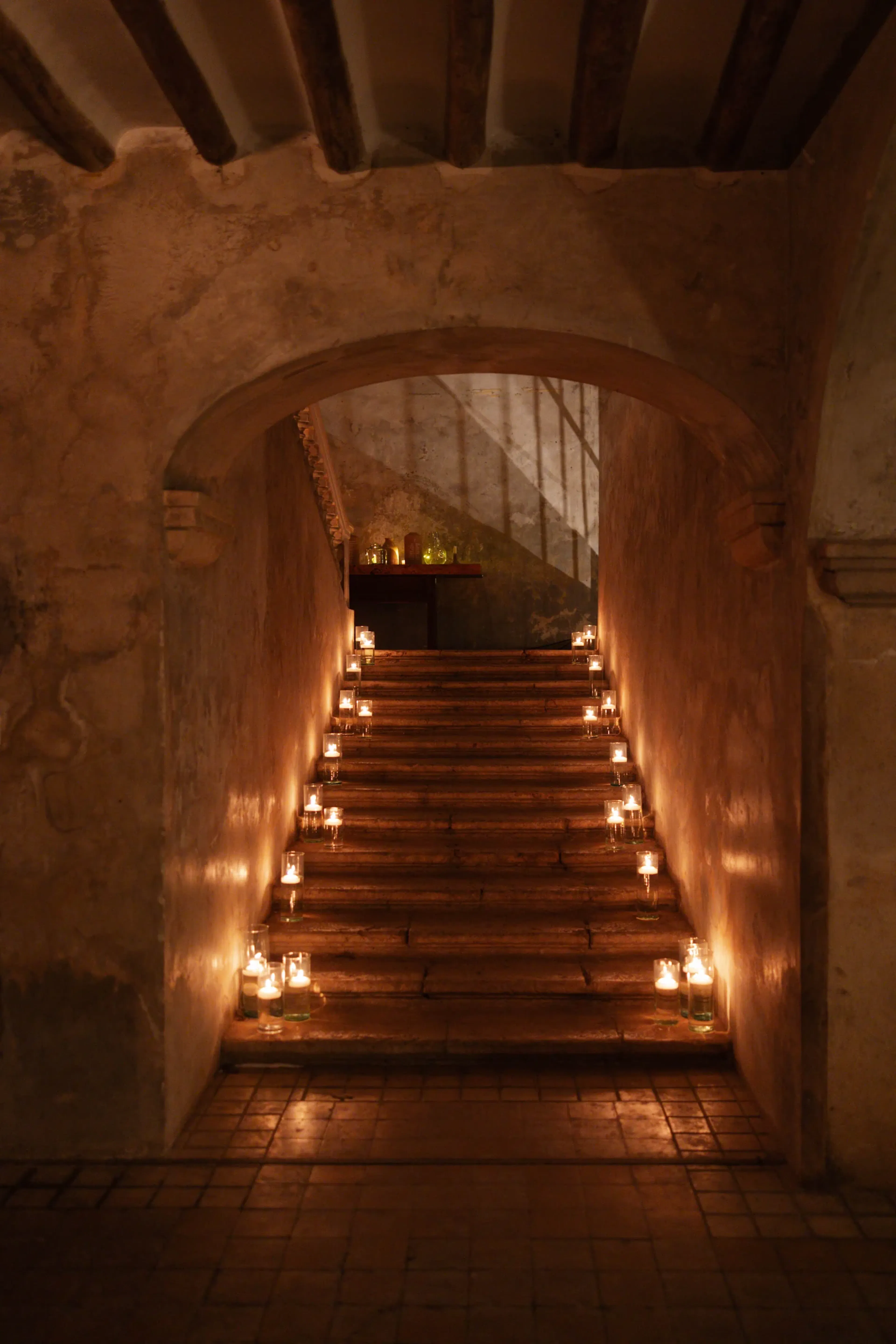 Una escalera de piedra tenuemente iluminada, adornada con velas en cada escalón, conduce a una puerta arqueada. La escena irradia un ambiente cálido y tranquilo.