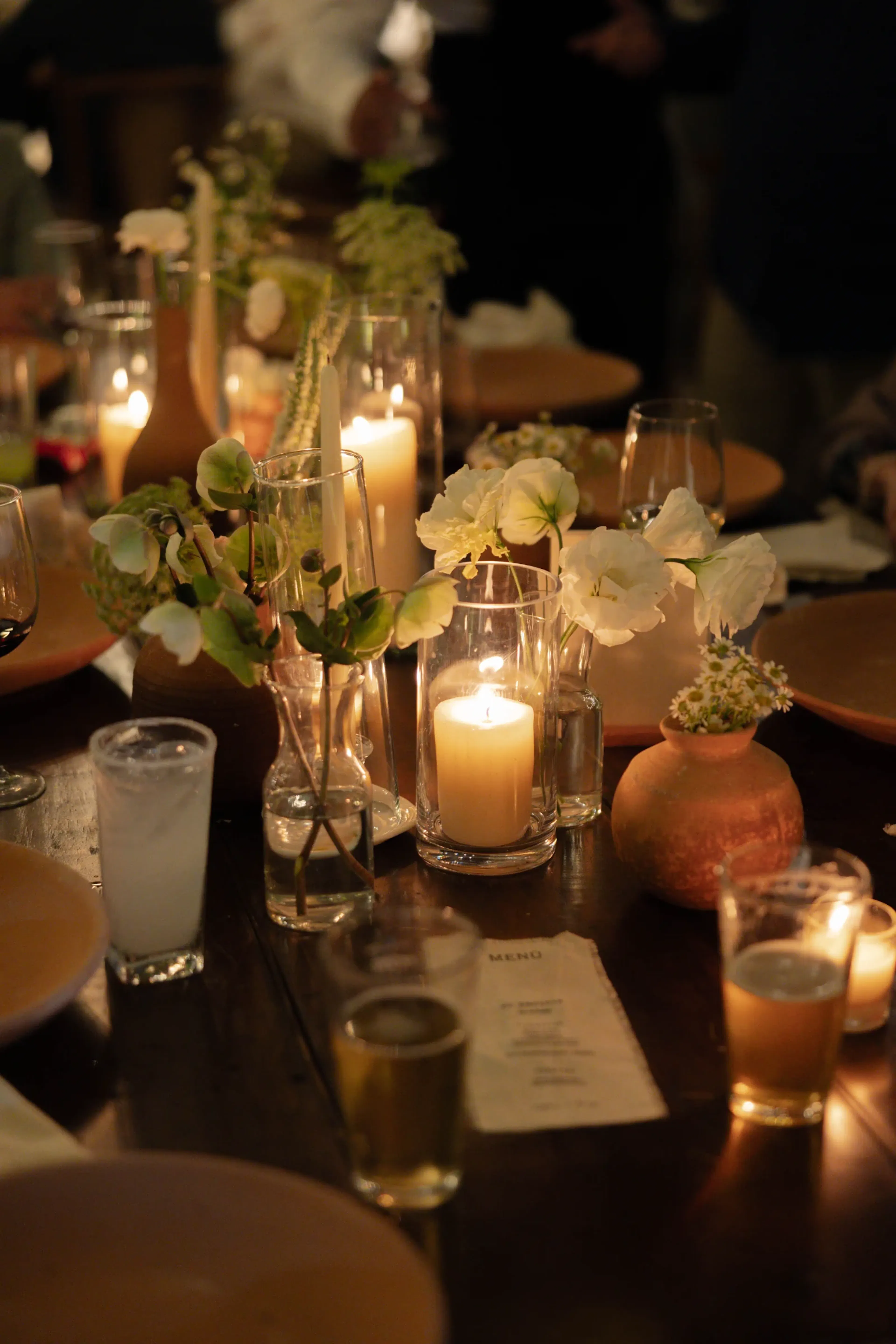 Elegante mesa de comedor con velas brillantes, flores blancas en jarrones, bebidas y un menú, creando un ambiente cálido e íntimo.