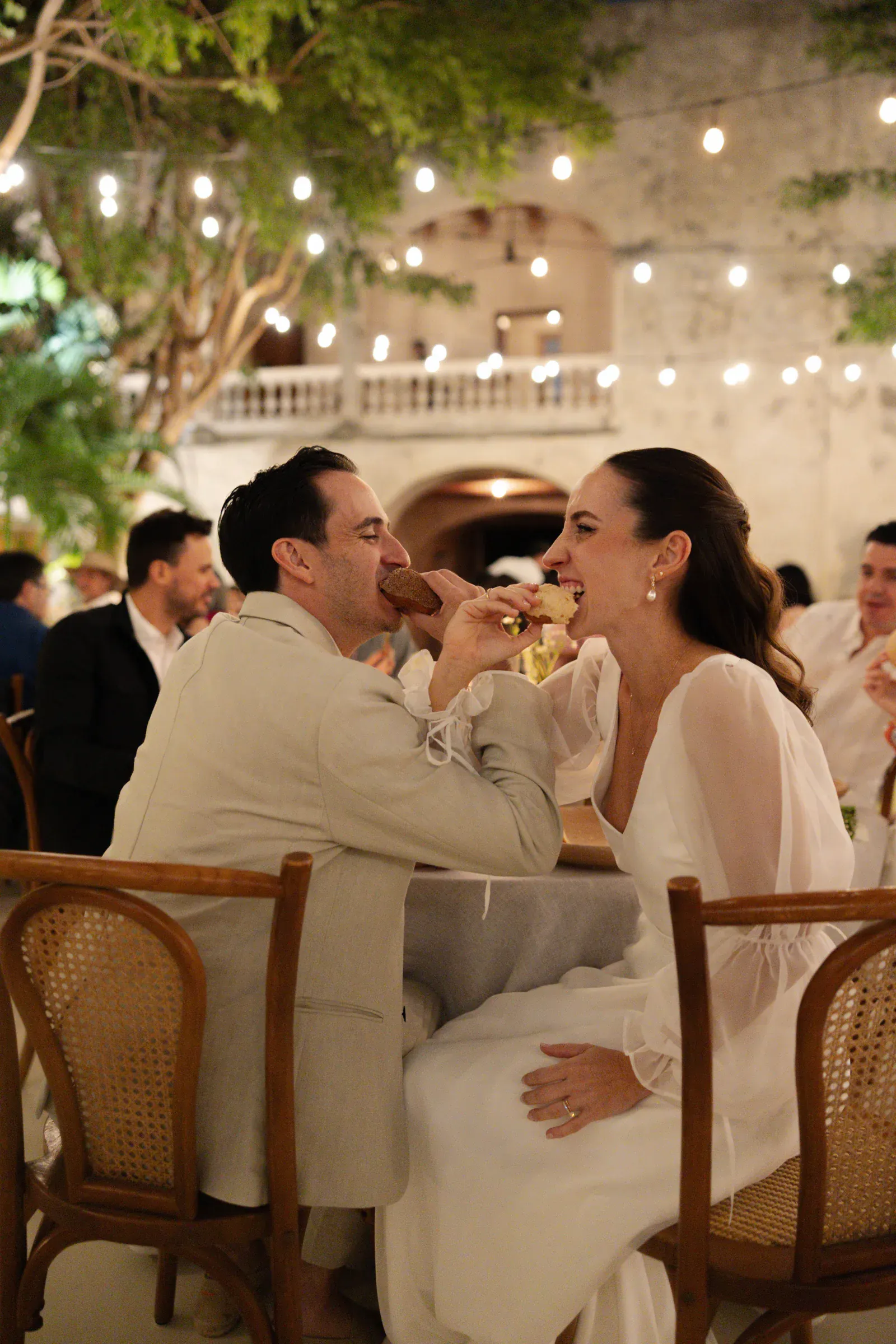 Una pareja, sonriente, se da de comer pastel alegremente en una velada al aire libre. Las guirnaldas de luces y los árboles crean un ambiente cálido y festivo a su alrededor.