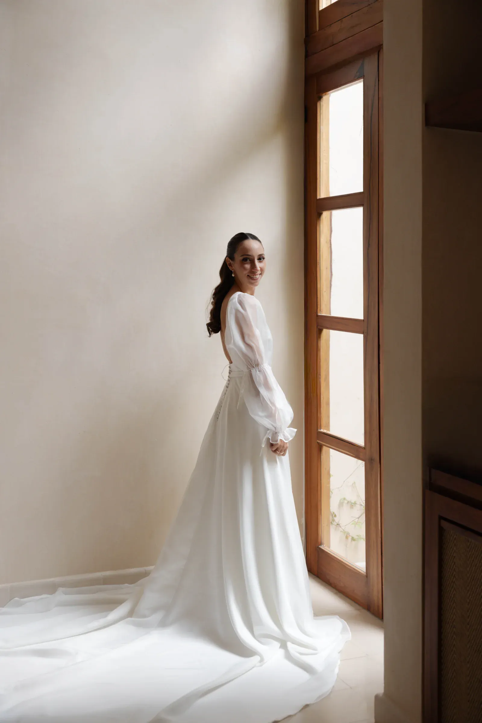 La novia, con un elegante vestido blanco, se encuentra junto a una gran ventanal, sonriendo suavemente. La luz natural realza la fluida cola y crea un ambiente sereno y alegre.