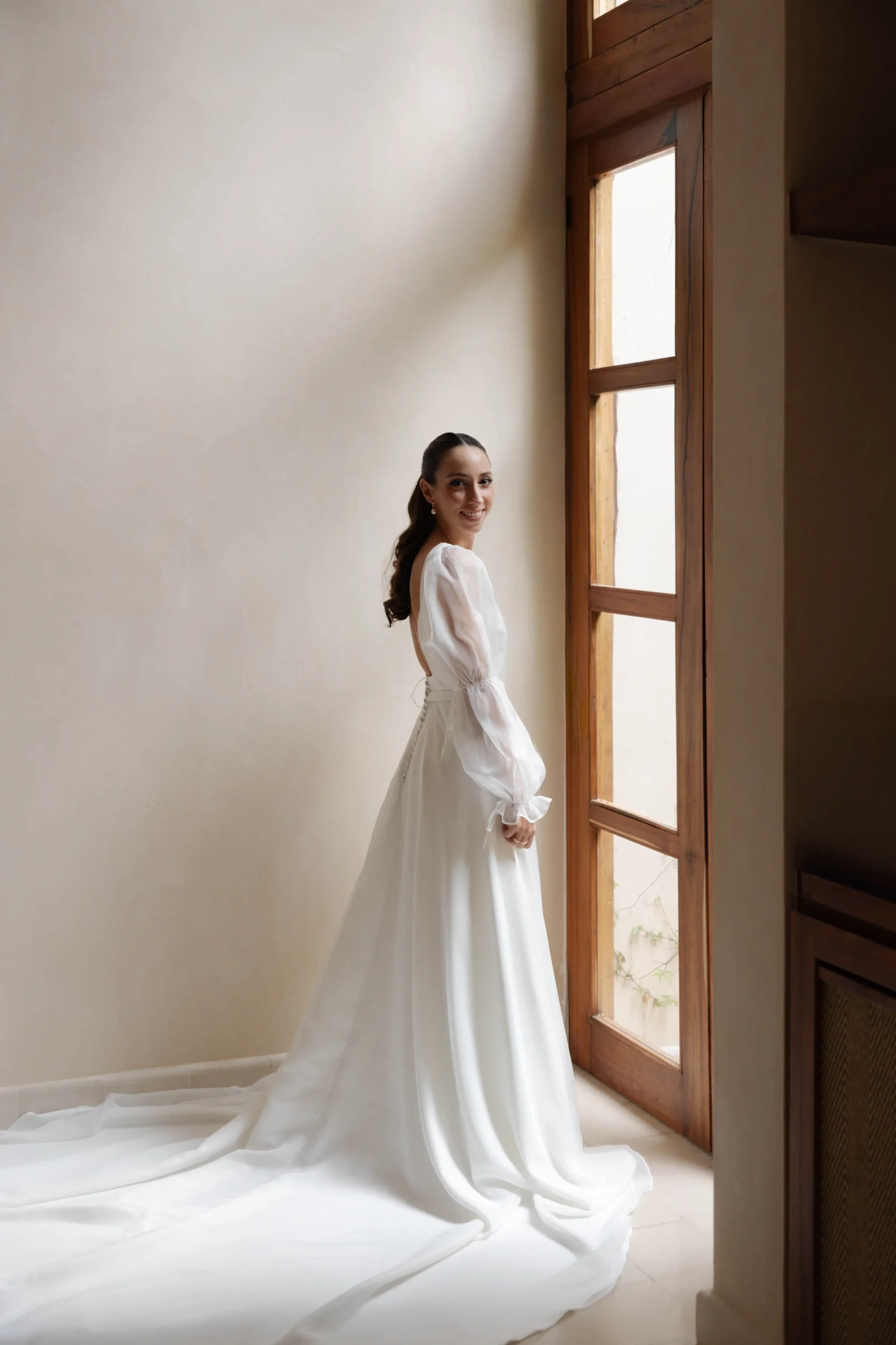 La novia, con un elegante vestido blanco, se encuentra junto a una gran ventanal, sonriendo suavemente. La luz natural realza la fluida cola y crea un ambiente sereno y alegre.