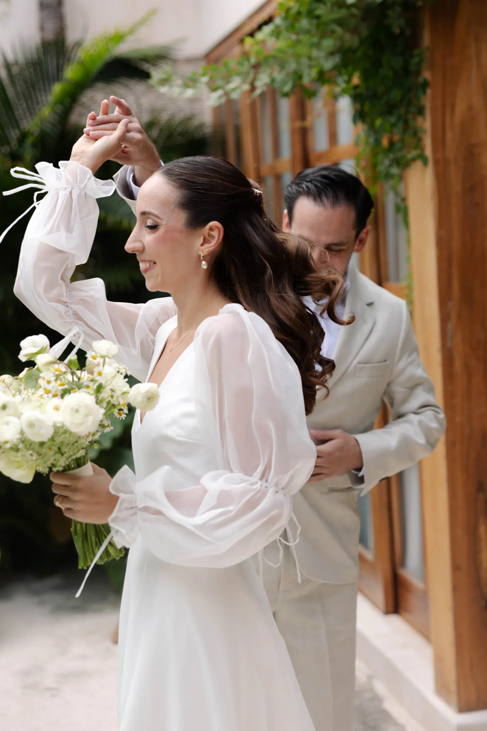La novia con un vestido blanco fluido sostiene un ramo y sonríe mientras baila con el novio con un traje beige, rodeado de vegetación y decoración de madera.