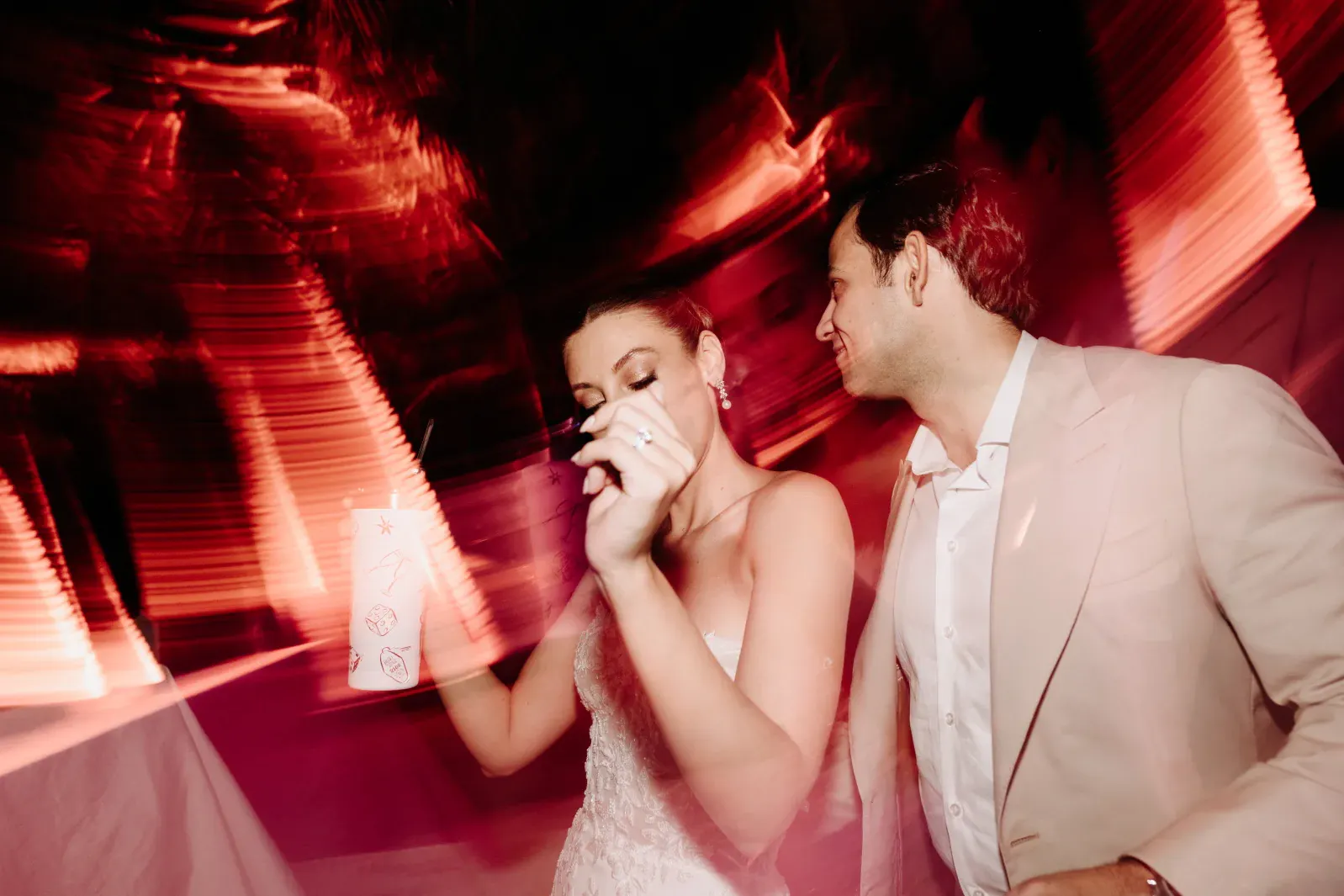 Una pareja baila alegremente bajo luces rojas y rosas que giran. La mujer, vestida de blanco, sostiene una bebida, mientras el hombre, vestido de traje beige, sonríe.