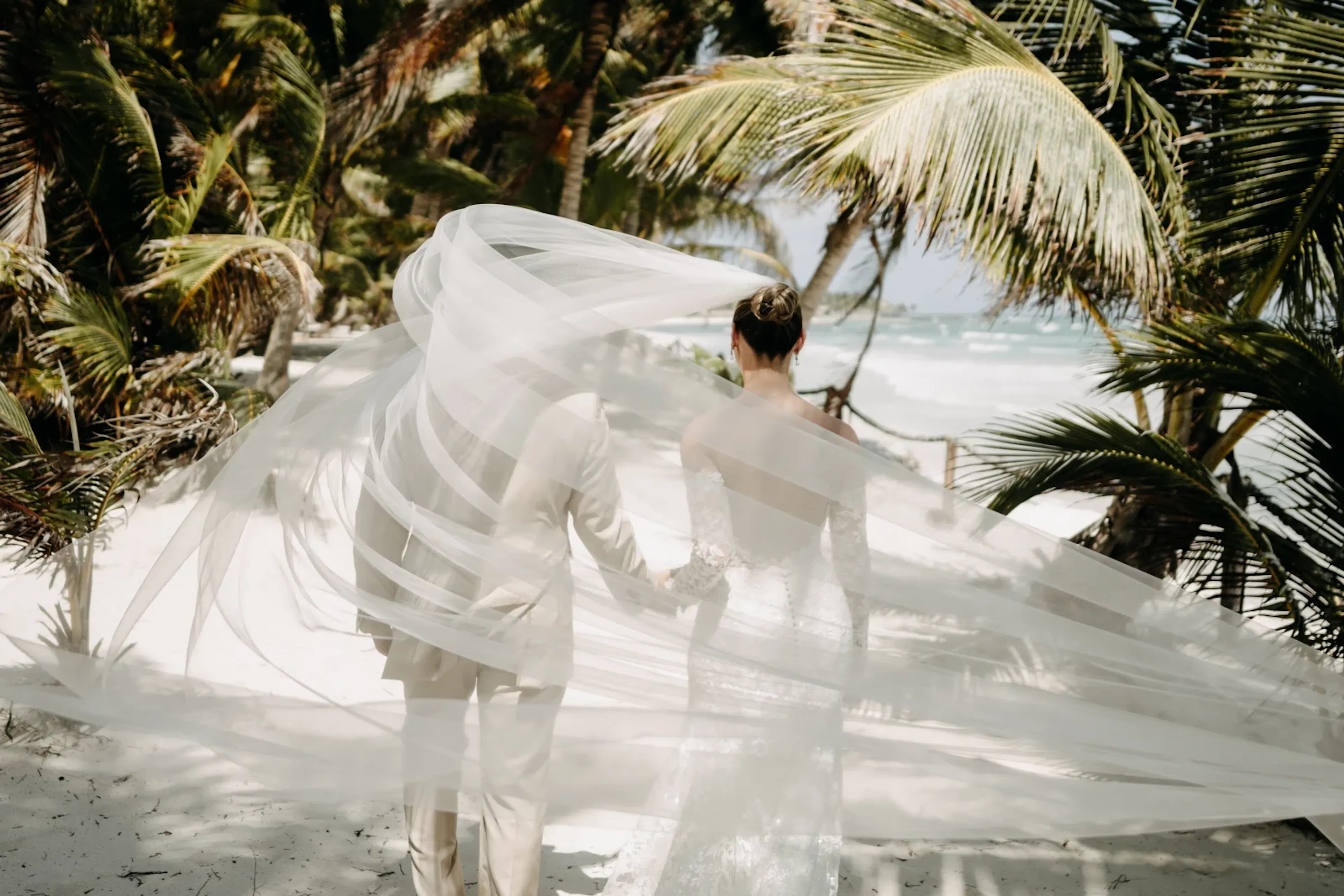 Una pareja con traje de novia camina por una playa tropical, entre palmeras meciéndose. Un velo ondea con la brisa, aportando elegancia y romanticismo.