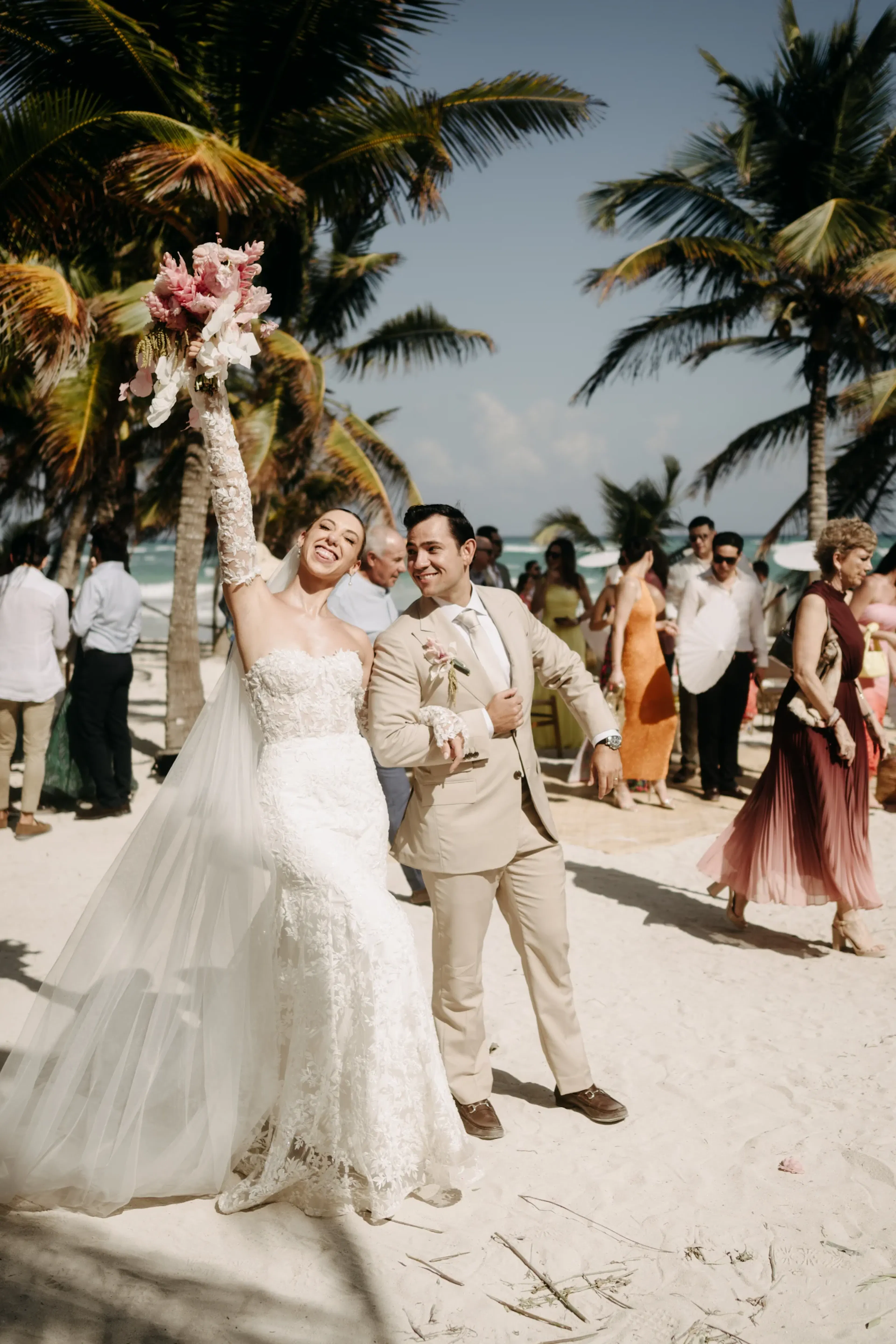Los novios posan alegremente en una playa soleada, rodeados de invitados. La novia sostiene un ramo de flores en alto, mientras las palmeras enmarcan la escena festiva.