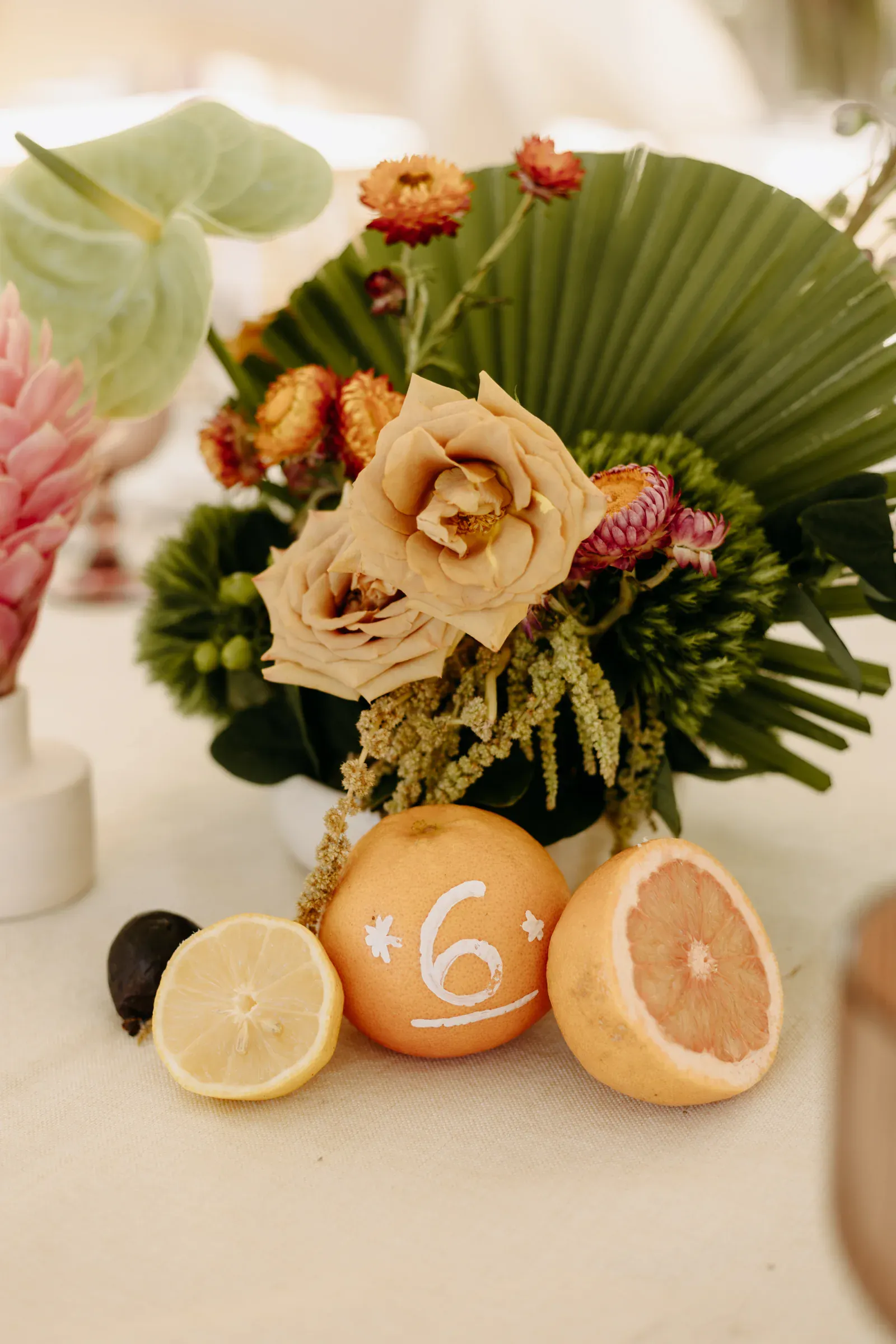 Un vibrante arreglo floral presenta rosas color melocotón, flores rosas y rojas, y grandes hojas verdes. Al frente, cítricos, con un pomelo marcado con un "6", que transmite una atmósfera alegre y veraniega.