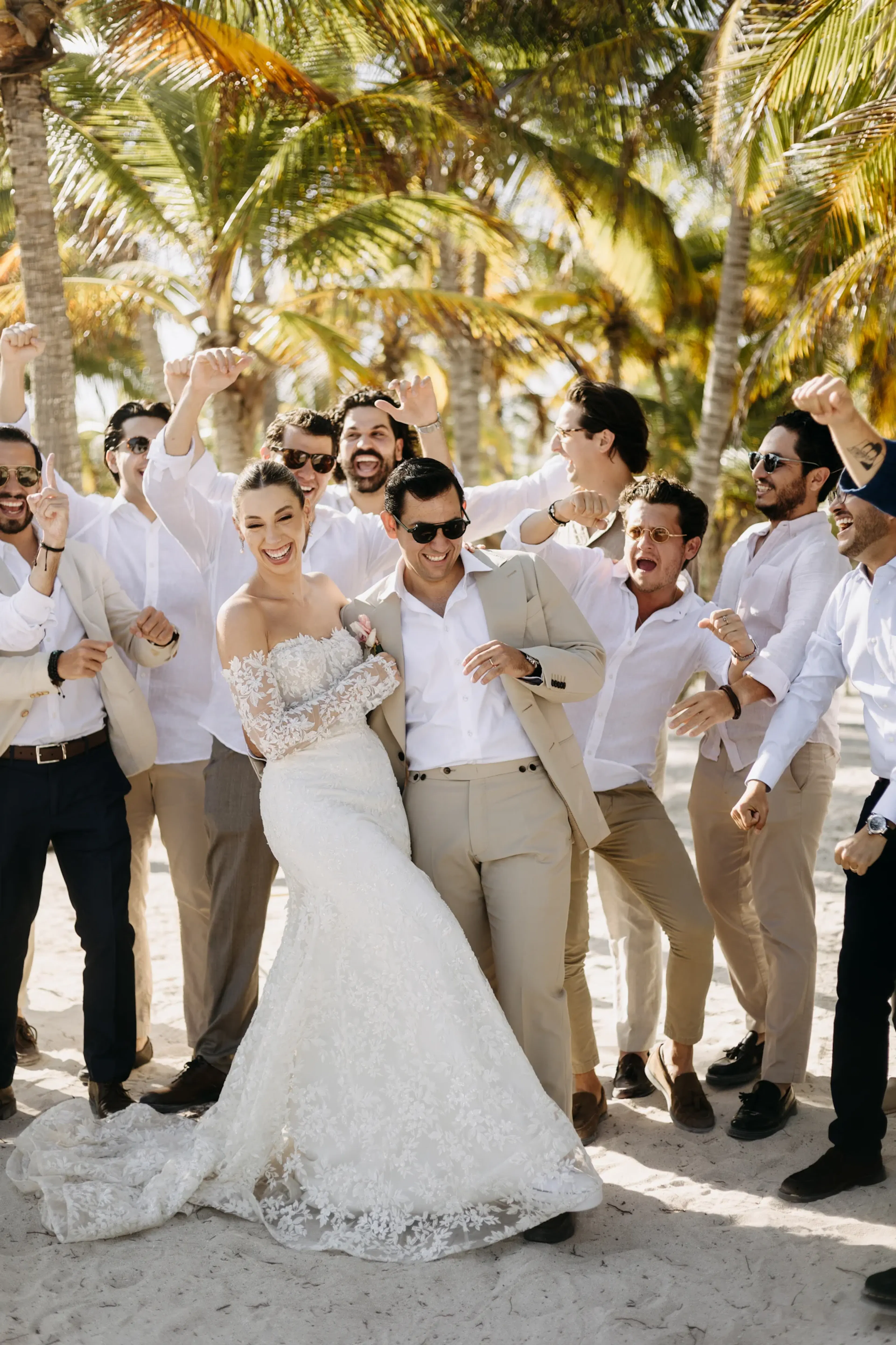 Los novios, elegantemente vestidos, sonriendo entre jubilosos padrinos de boda con trajes y camisas claras, posan bajo palmeras iluminadas por el sol en una playa de arena.