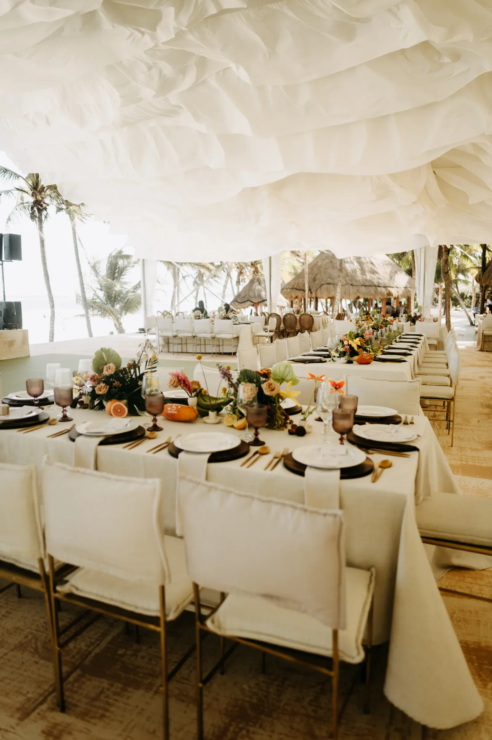 Elegante comedor junto a la playa con largas mesas adornadas con flores, platos y cristalería bajo un ondulante dosel blanco, que irradia un ambiente sereno y festivo.
