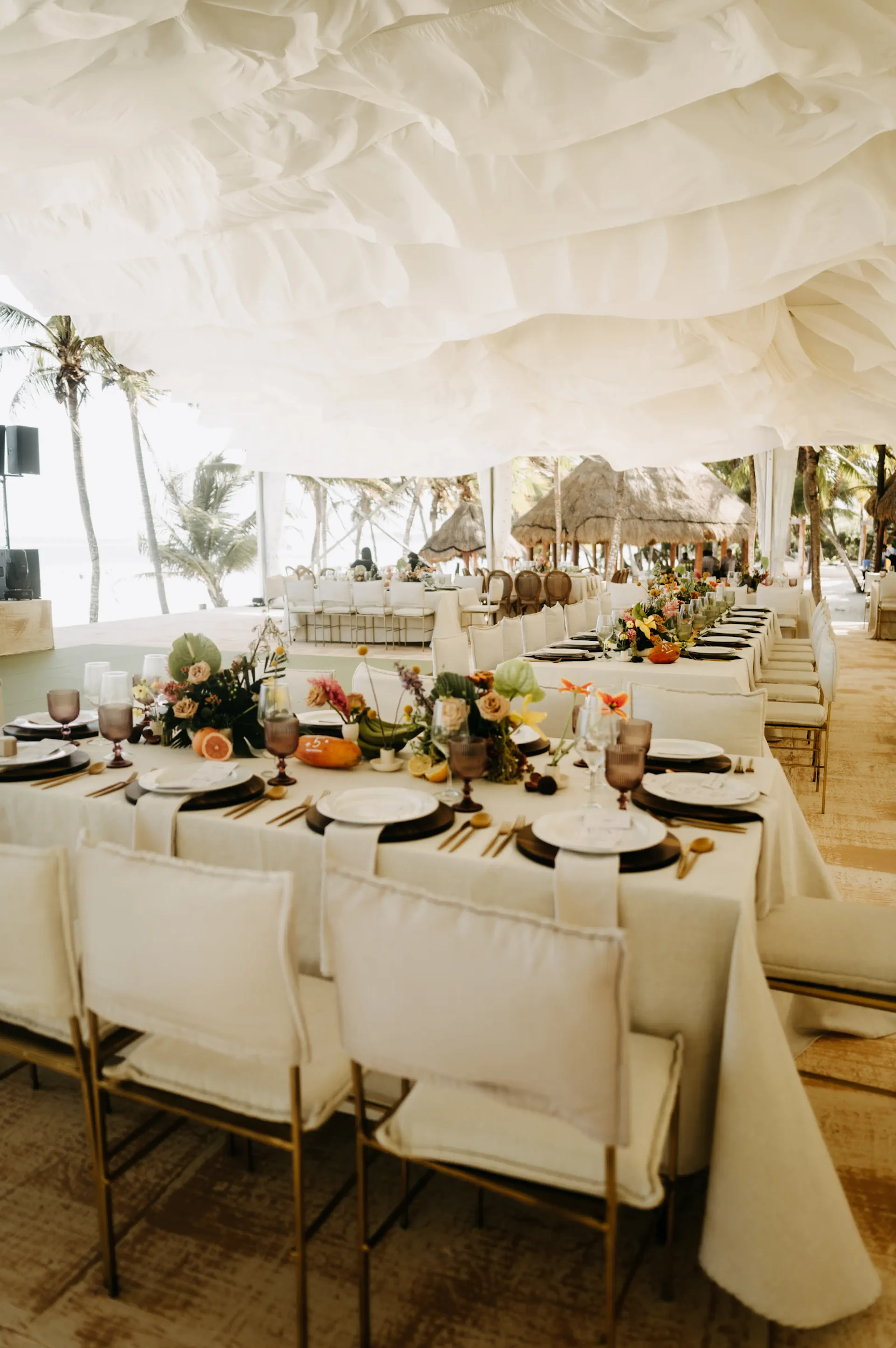 Elegante comedor junto a la playa con largas mesas adornadas con flores, platos y cristalería bajo un ondulante dosel blanco, que irradia un ambiente sereno y festivo.