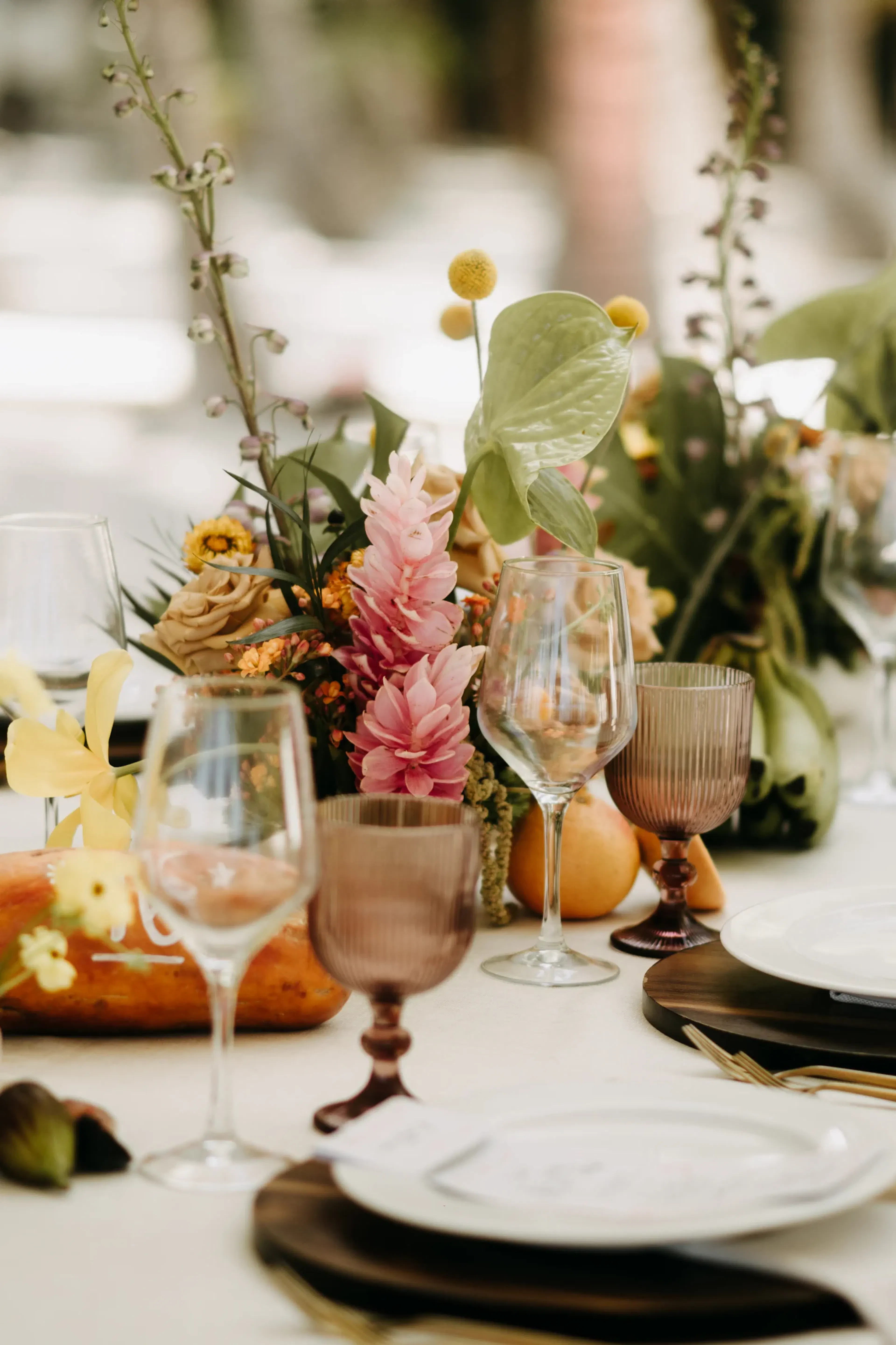 Elegante mesa con copas y platos variados sobre un mantel blanco. Un exuberante centro de mesa floral, con flores rosas y amarillas, le da un toque vibrante.