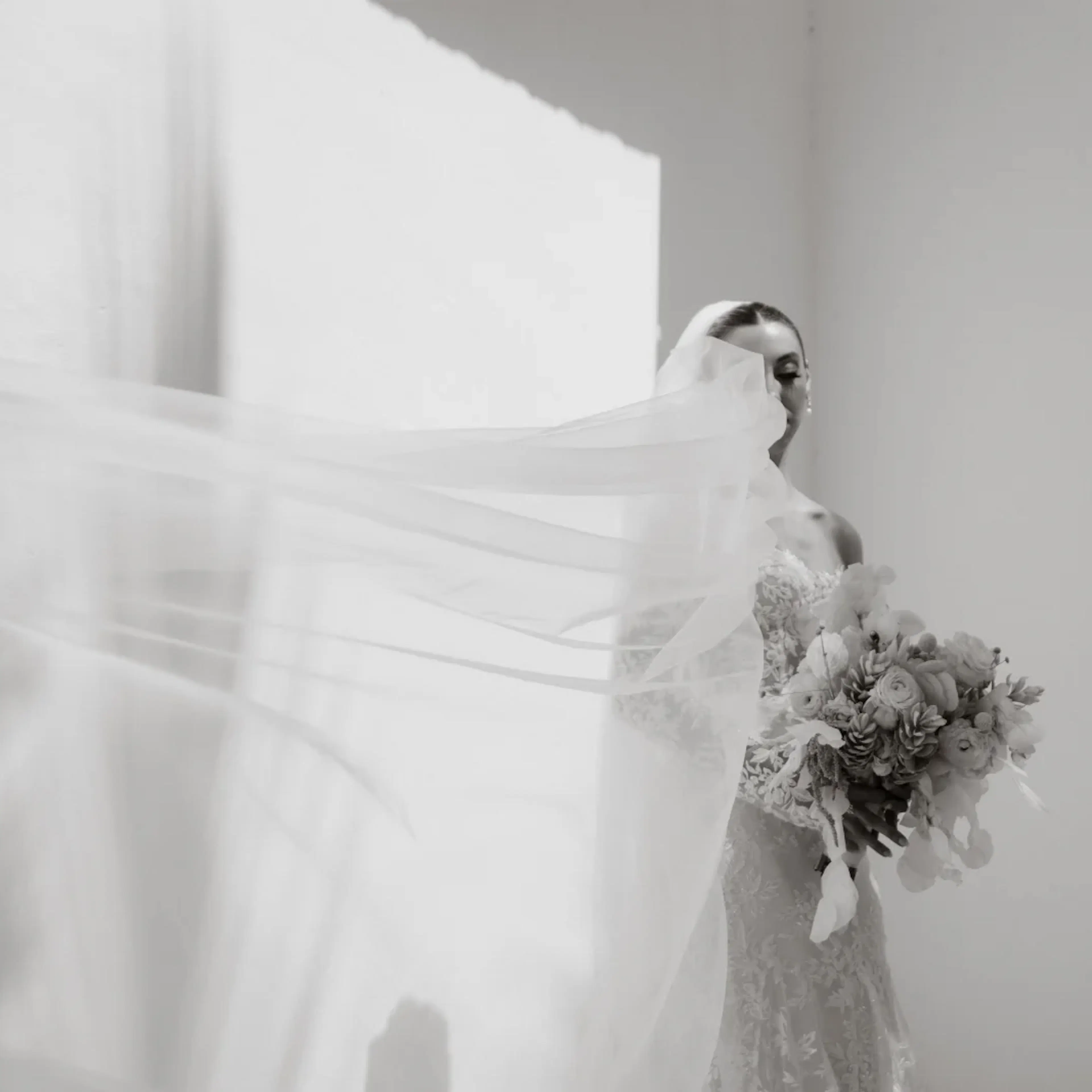 Fotografía en blanco y negro de una novia con un delicado vestido de encaje, sosteniendo un ramo. Su velo ondea con gracia bajo la suave luz, creando una atmósfera serena y etérea.