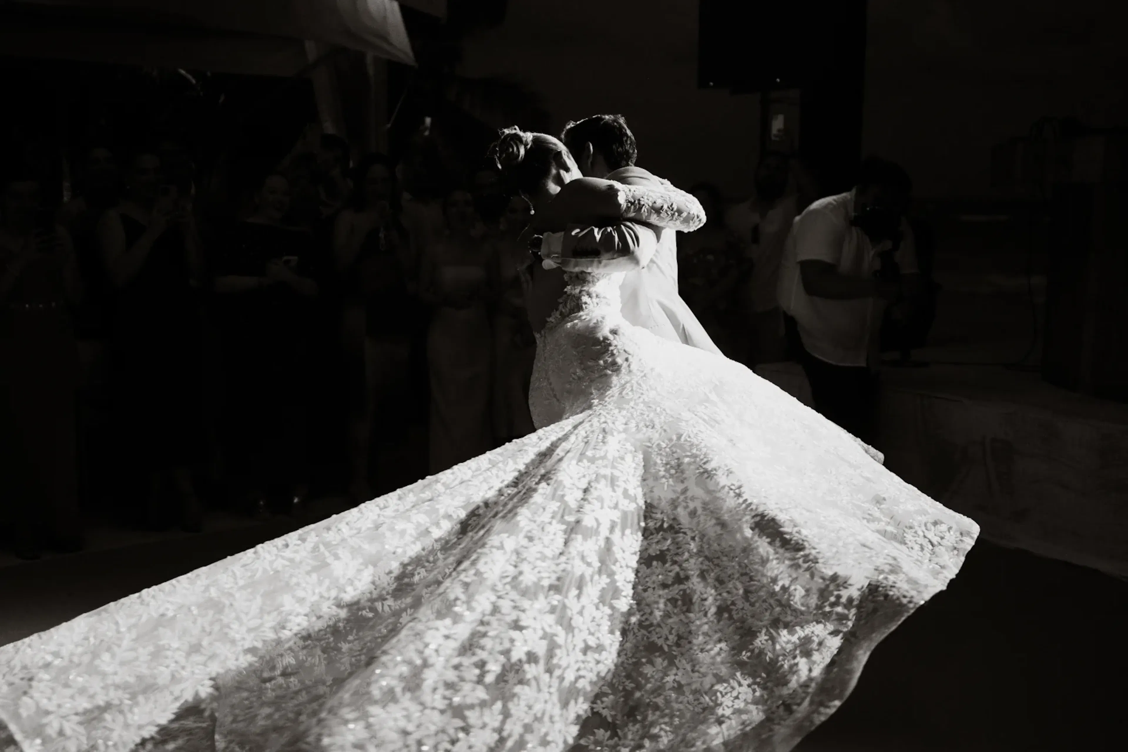 Fotografía en blanco y negro de una novia con un delicado vestido de encaje, bailando con el novio. Su vestido ondea con gracia bajo la suave luz, creando una atmósfera romántica y etérea.