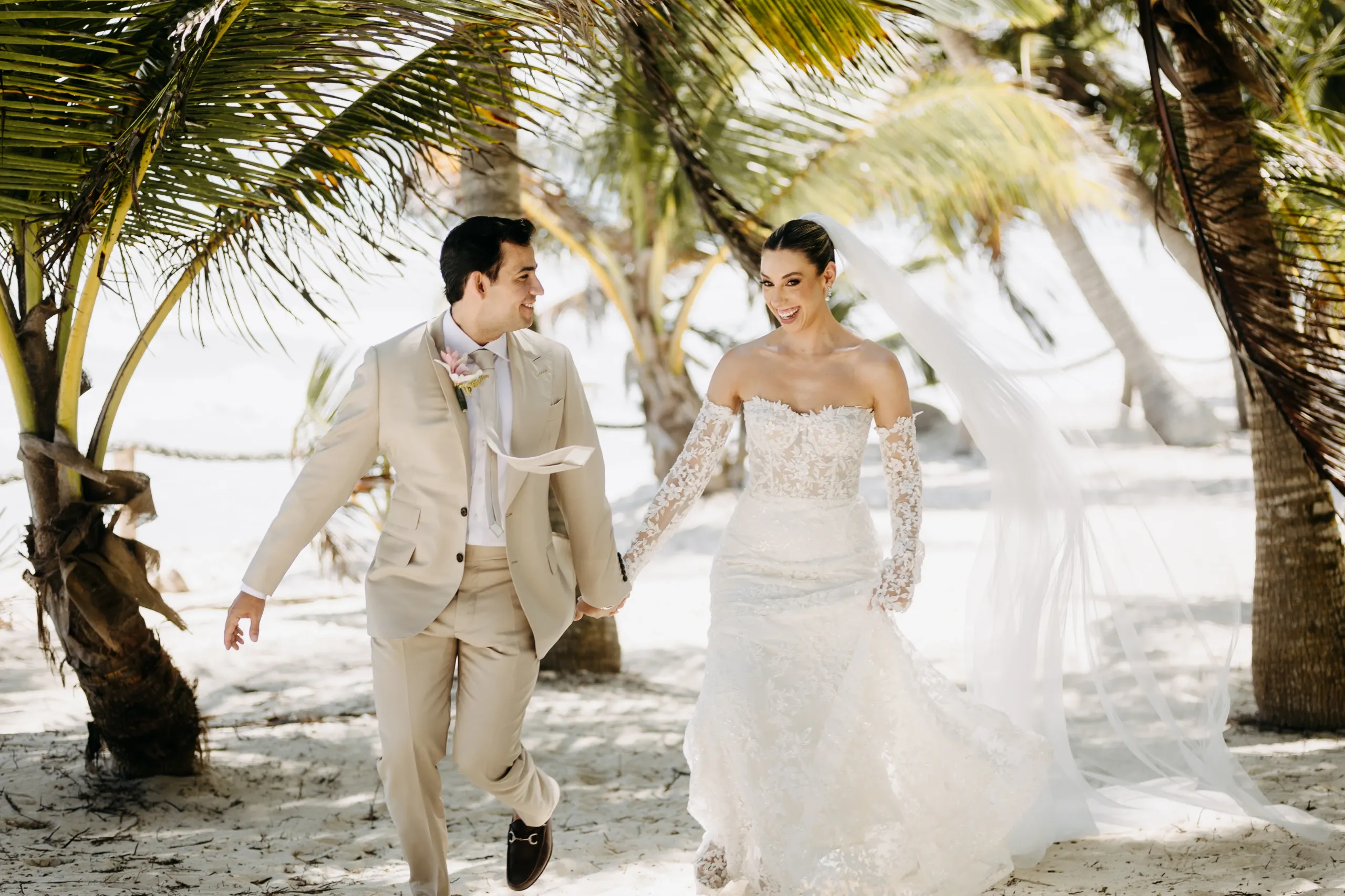 Una pareja alegre, la novia con un vestido de encaje y velo, el novio con un traje beige, caminan de la mano bajo palmeras en una playa iluminada por el sol.