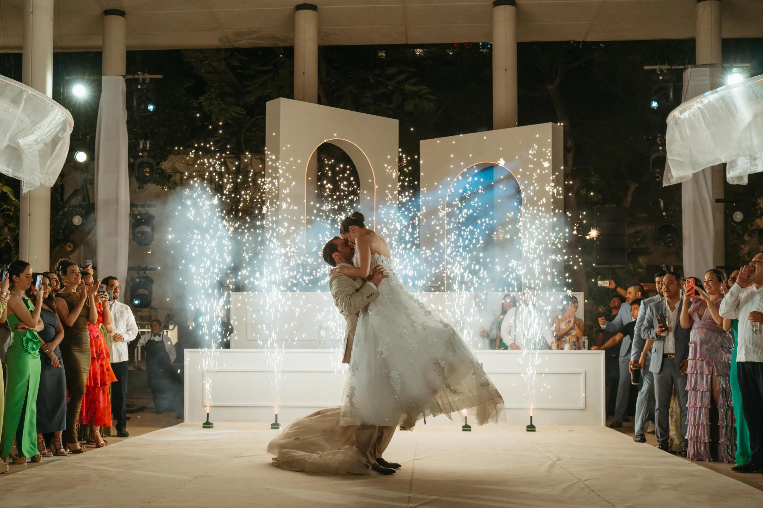 Una pareja de recién casados comparte un alegre baile en medio de brillantes fuegos artificiales en un escenario brillante y elegante, rodeados de invitados que aplauden con atuendos vibrantes.