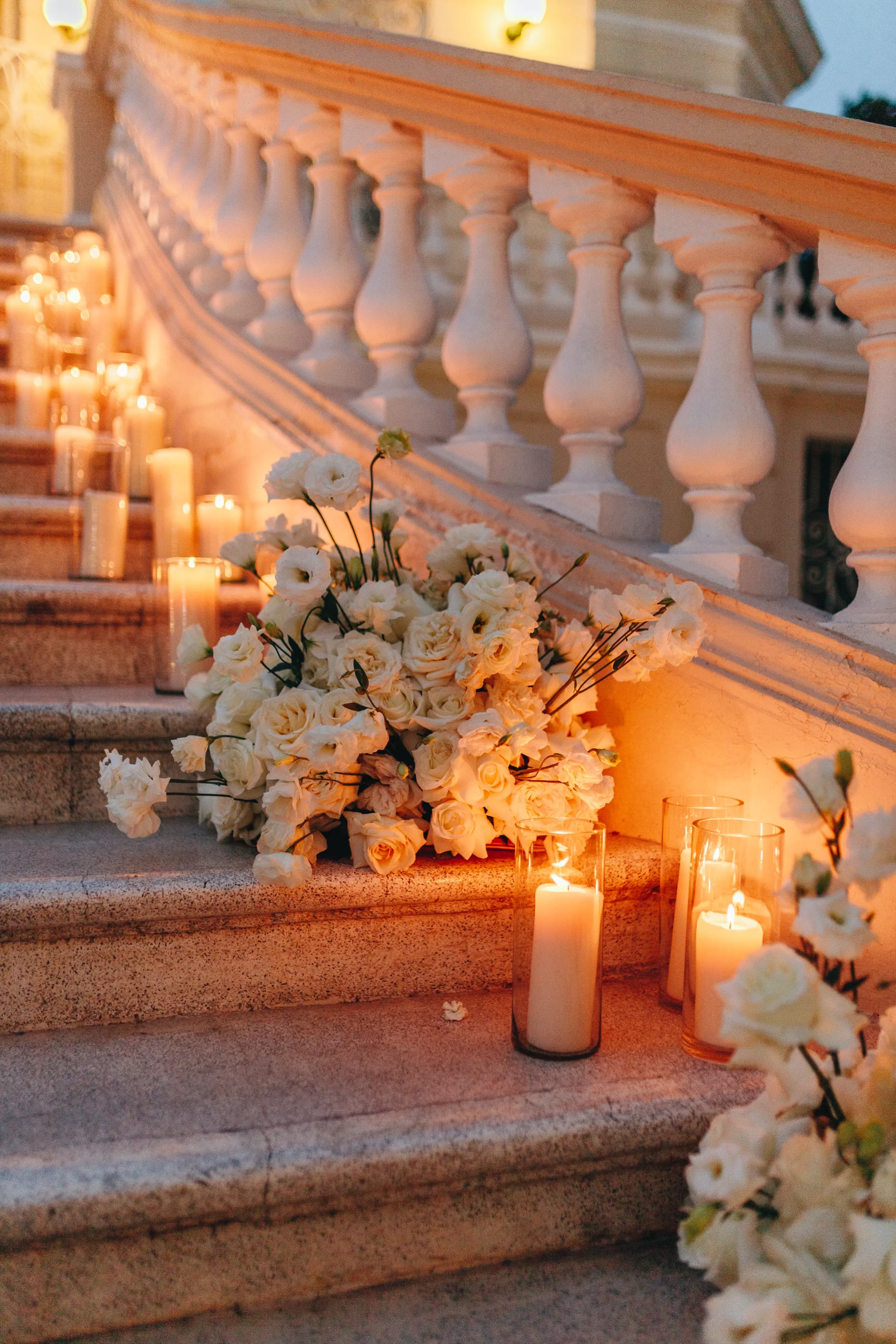 Escalera romántica adornada con velas brillantes y ramos de rosas blancas, creando un ambiente sereno y elegante bajo la suave luz del atardecer.