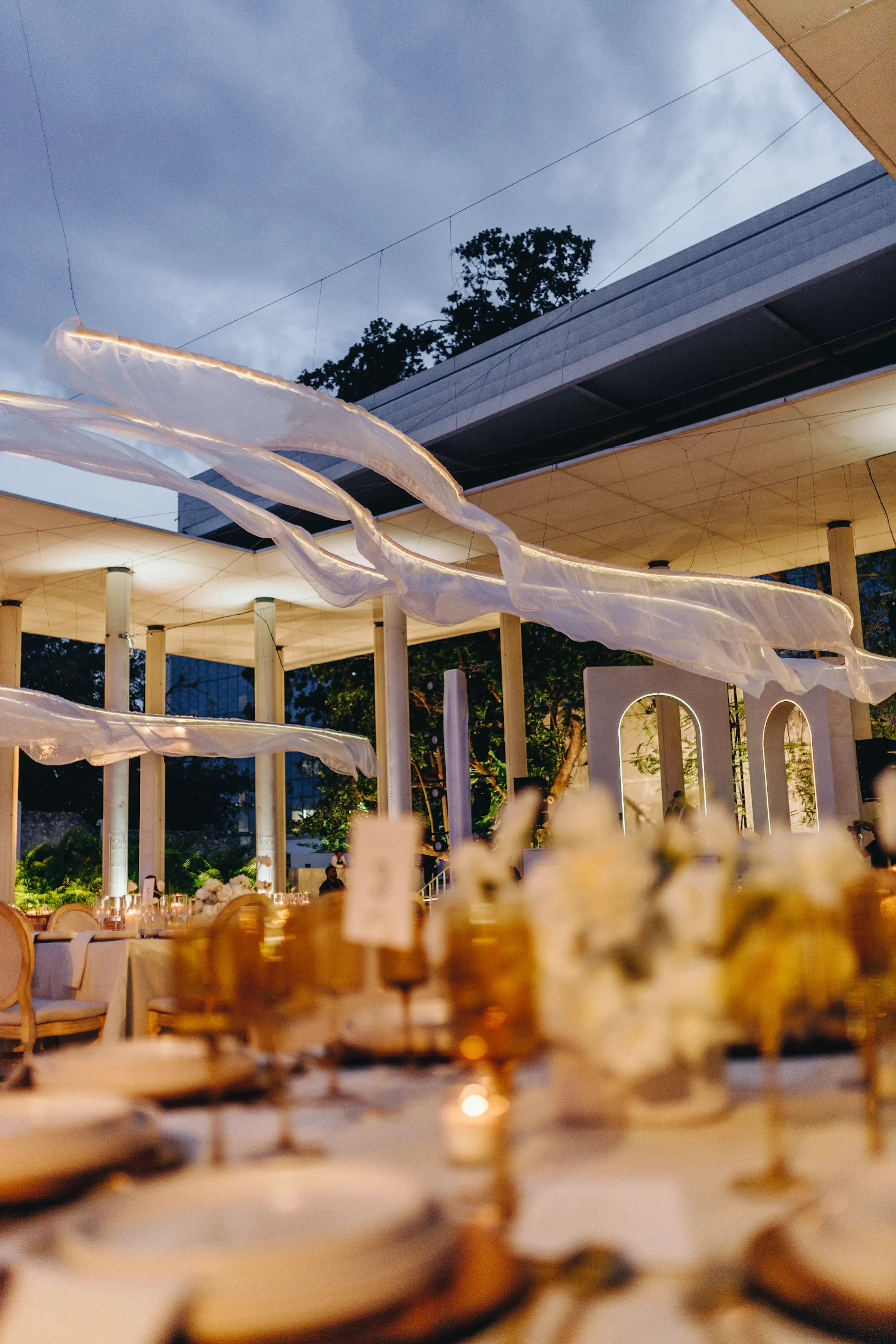 Elegante evento nocturno al aire libre con mesas preparadas para cenar, adornadas con velas y detalles dorados. Telas transparentes cubren el techo, creando un ambiente sofisticado.