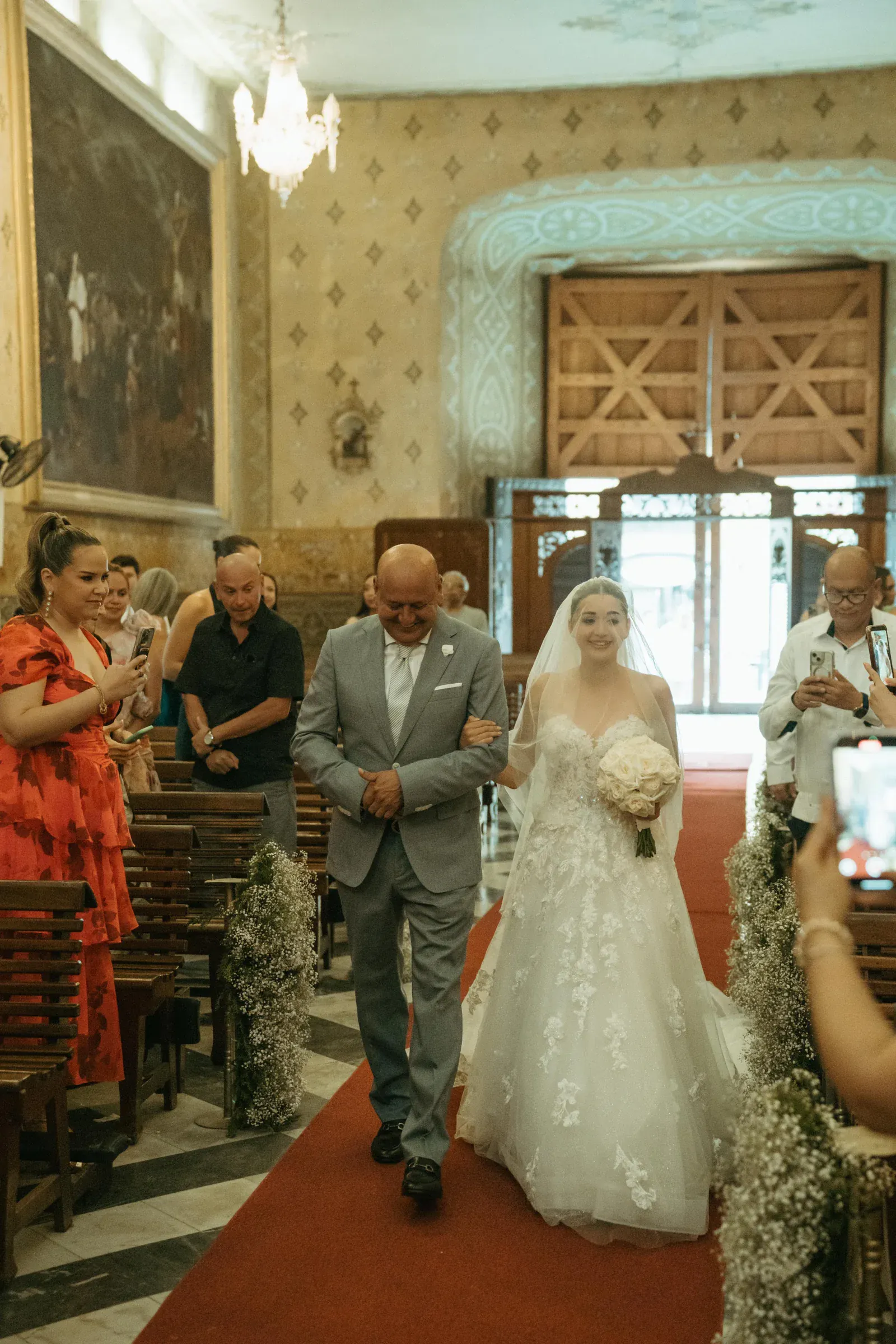 Una novia con un vestido blanco sonríe mientras camina del brazo de un hombre mayor por el pasillo de una iglesia alfombrada de rojo. Los invitados a ambos lados capturan el momento.