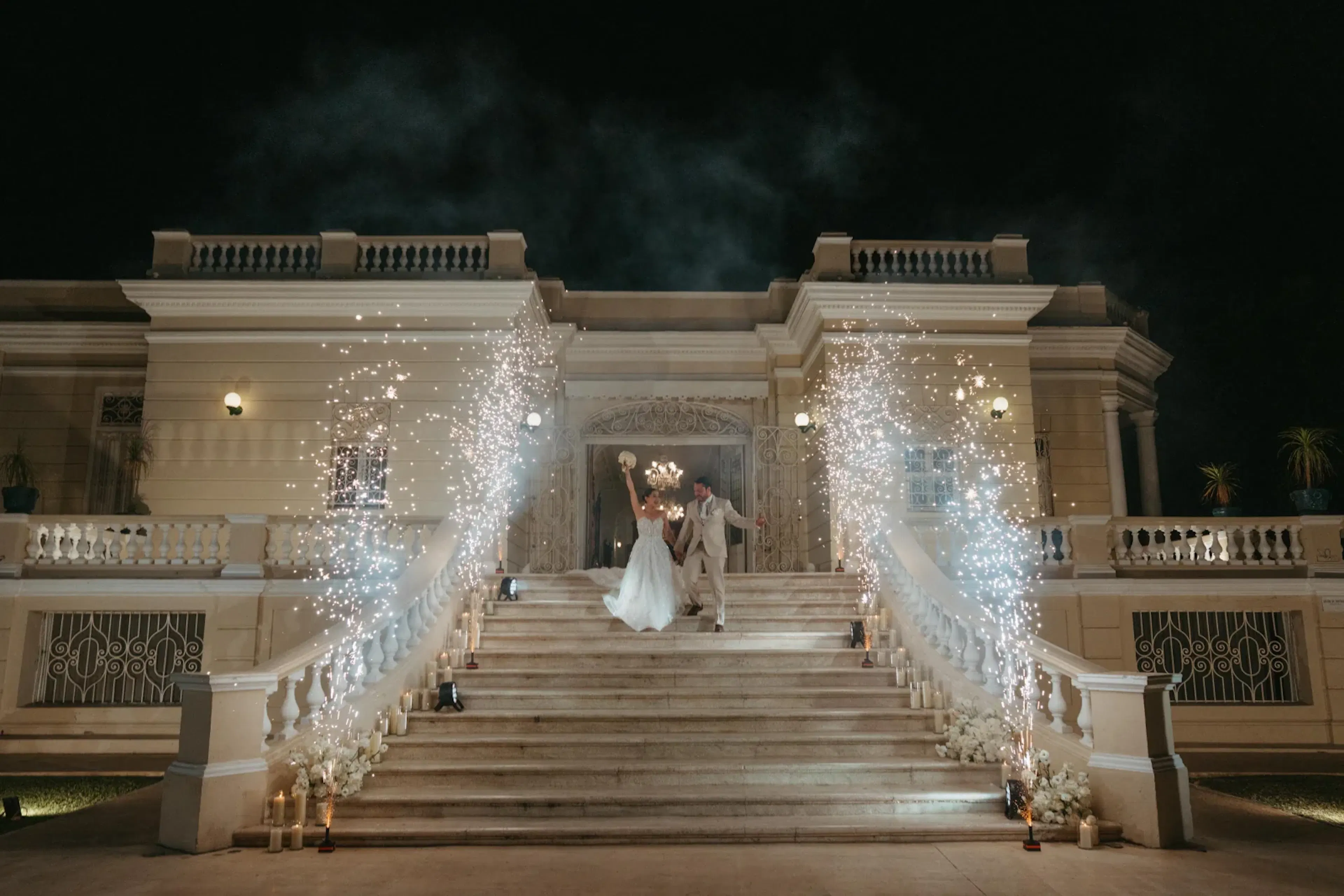 Una pareja, vestida de novia, se encuentra en una gran escalinata, tomada de la mano bajo el cielo nocturno. Los fuegos artificiales iluminan su entrada, creando un ambiente festivo y romántico.
