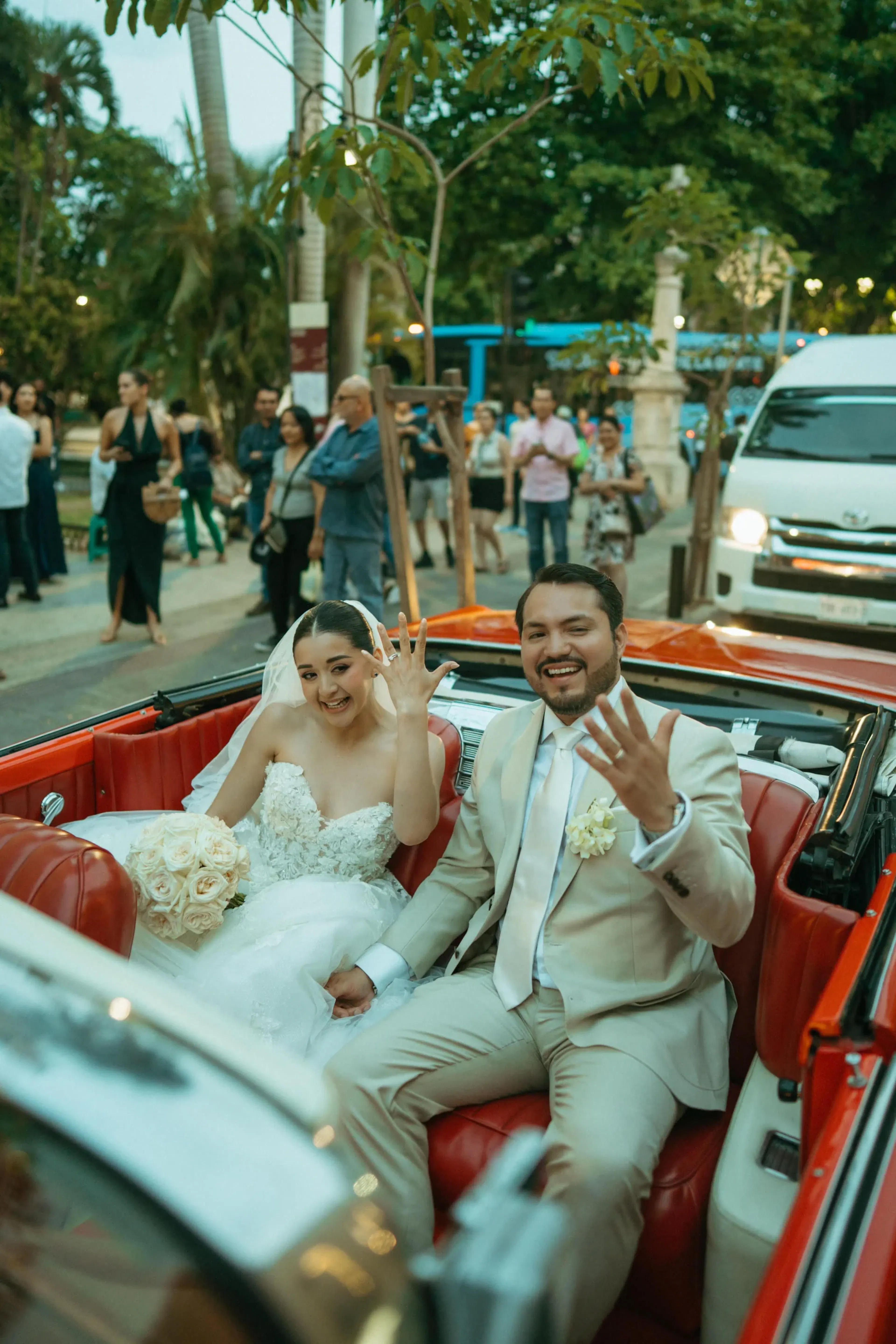 Los novios muestran alegremente sus anillos sentados en un descapotable rojo. La novia sostiene un ramo de flores; los espectadores vitorean al fondo.
