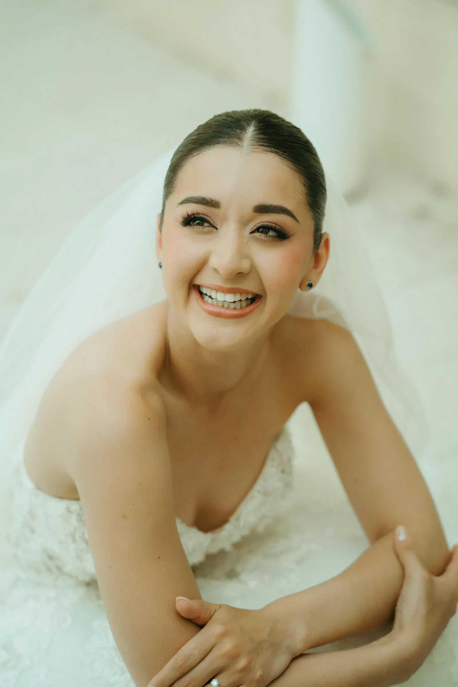 Novia sonriente con velo y vestido palabra de honor, recostada sobre una superficie blanca. Su expresión alegre crea una atmósfera de felicidad y celebración.