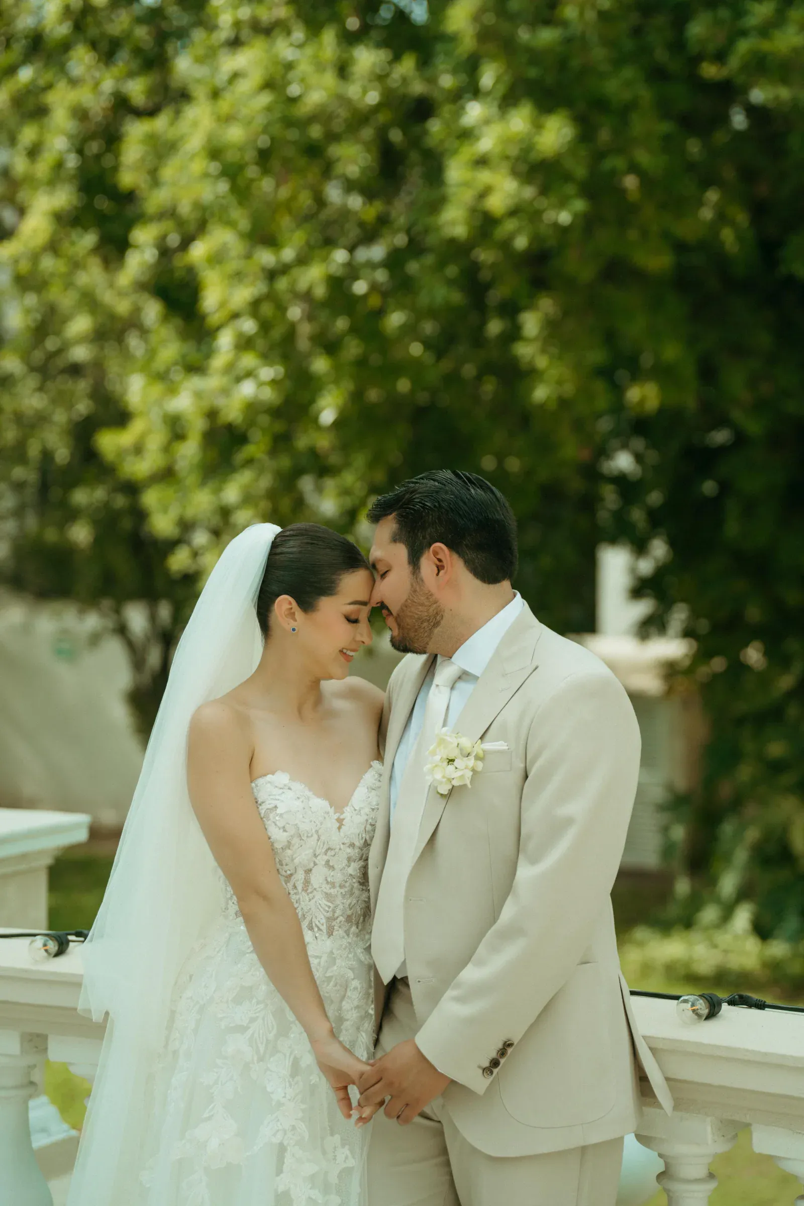 Los novios, vestidos de gala, comparten un momento de ternura, con las frentes tocándose y sonriendo, frente a un exuberante jardín verde como telón de fondo. Romántico y sereno.